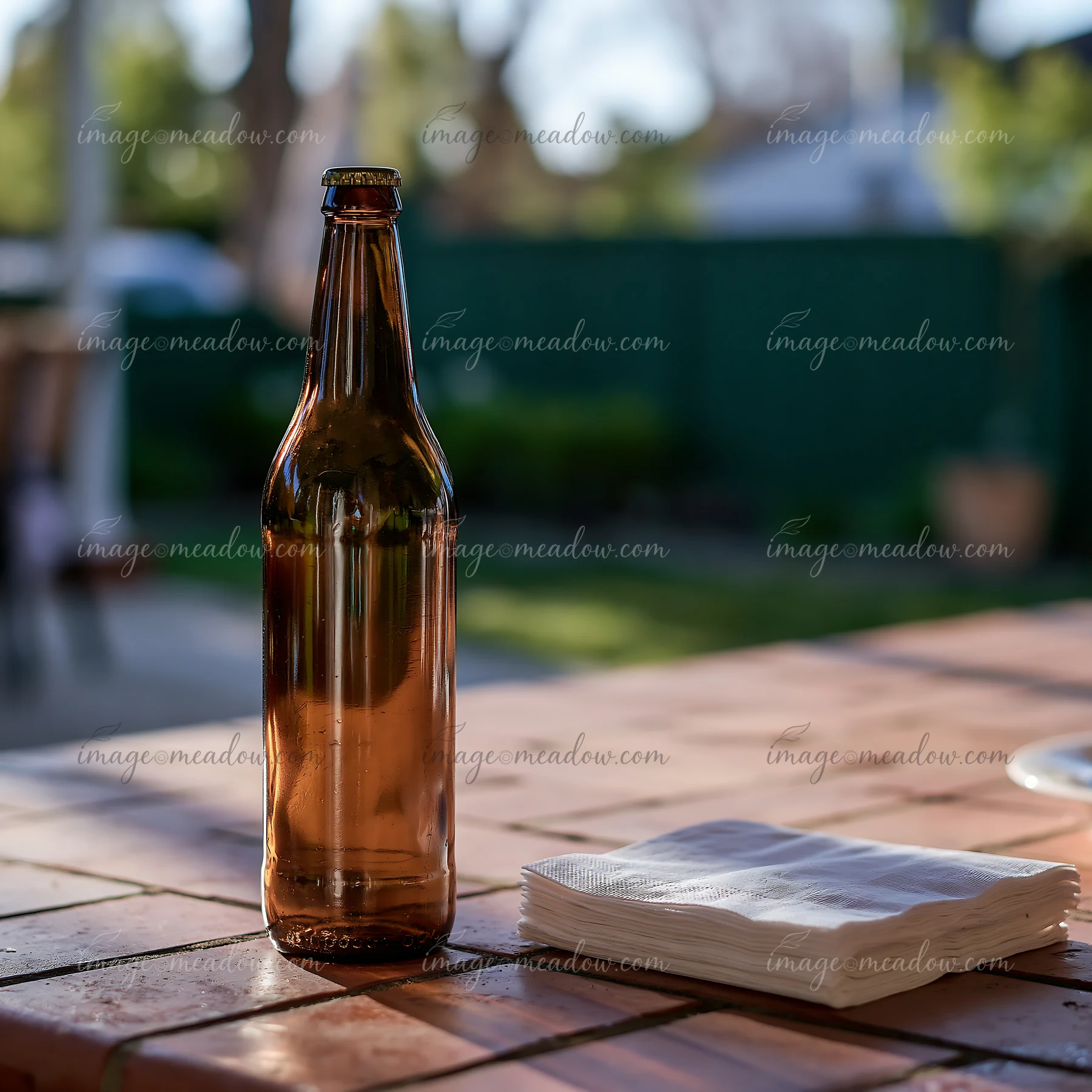 An empty bottle sits on a wooden table beside a stack of napkins at a backyard gathering during the day.