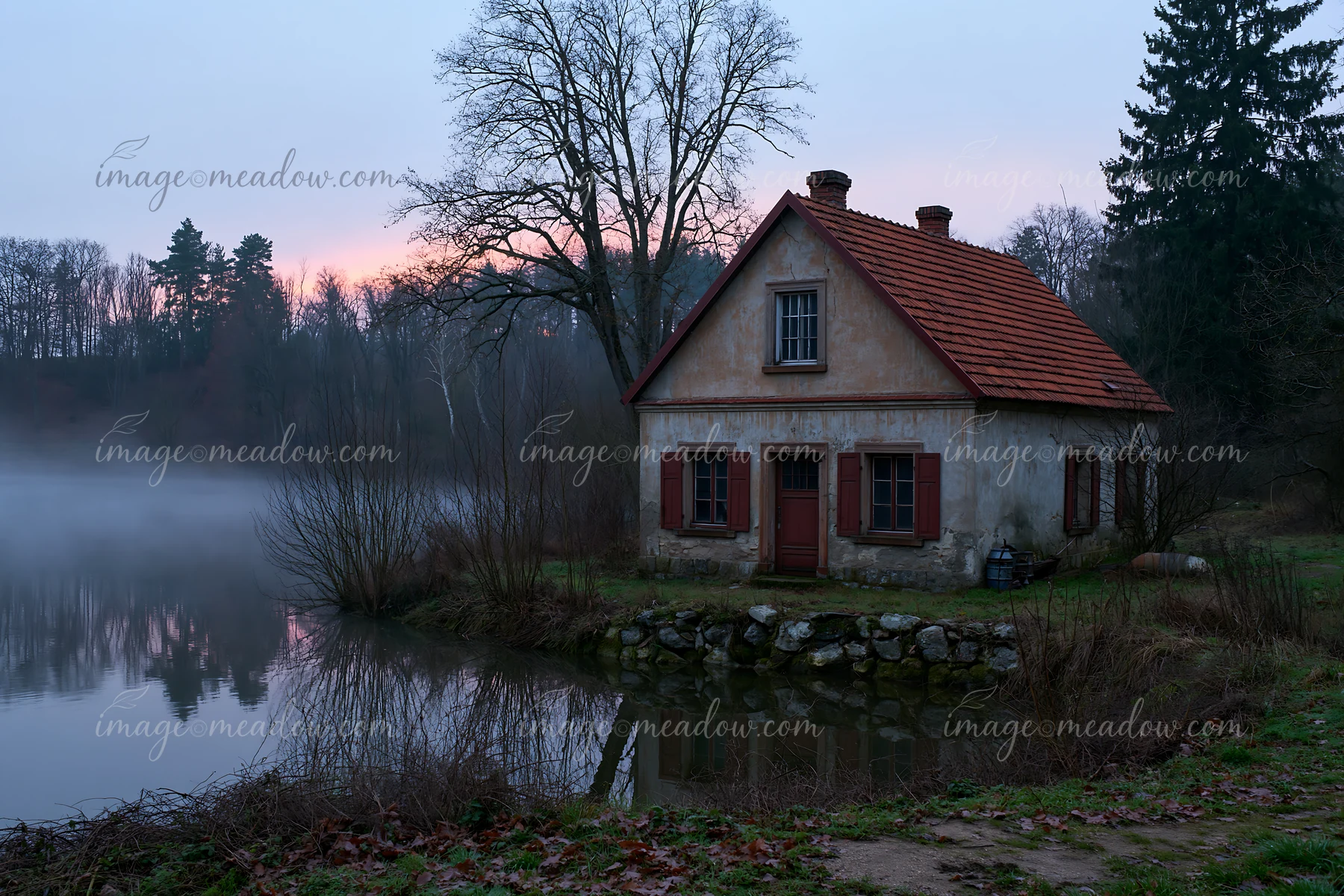 An old cottage with weathered facade stands beside a calm lake surrounded by trees.