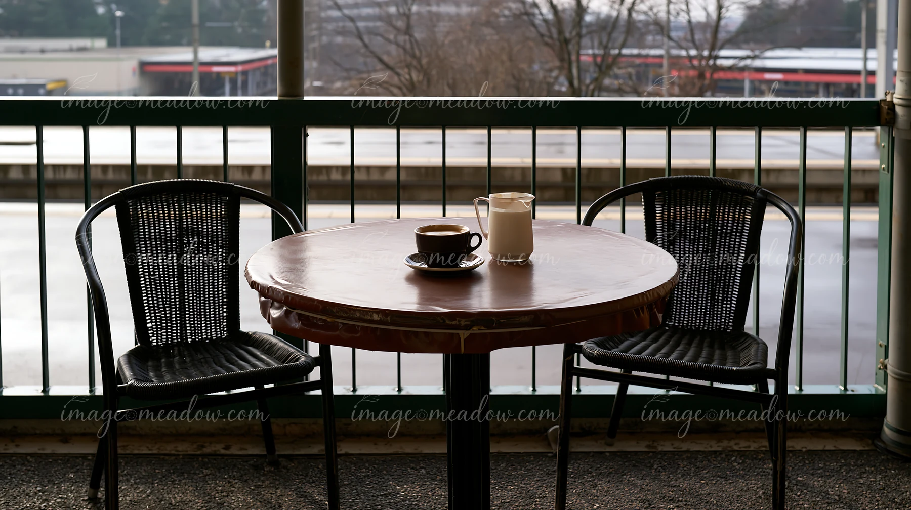 An outdoor cafe table holds a latte and a milk jug with two black chairs nearby.