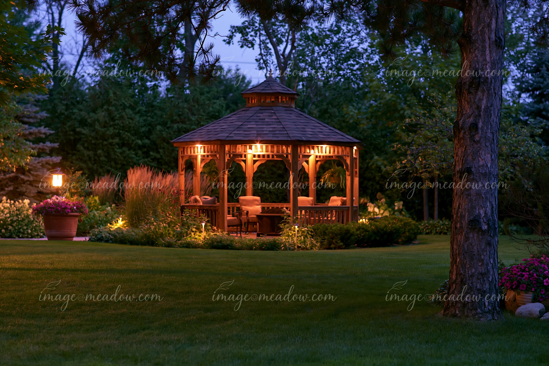 An upscale garden pavilion provides a cozy seating area surrounded by a well-kept lawn and lush greenery at dusk.