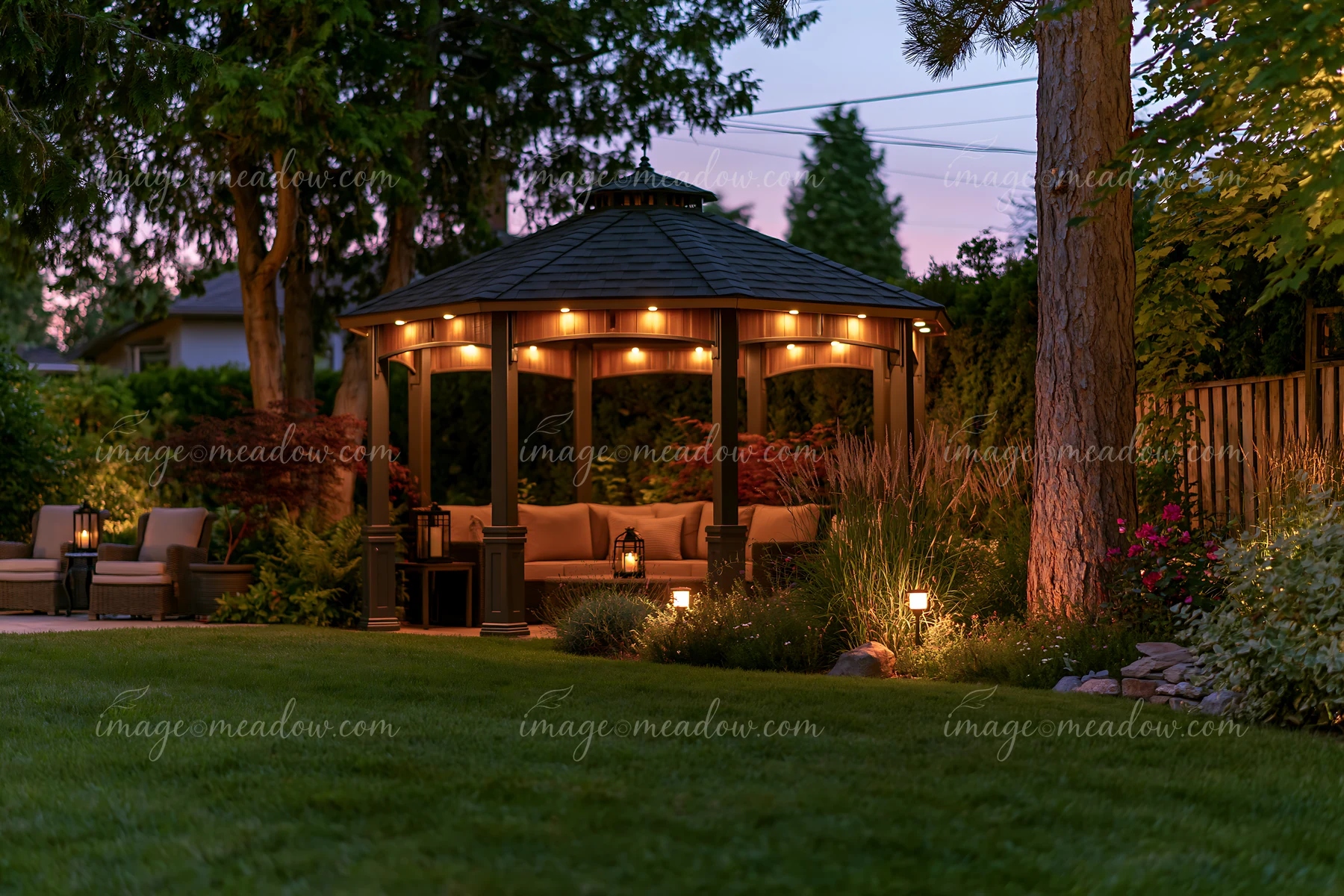 An upscale garden pavilion with lights stands beside a lawn at dusk, surrounded by trees and plants.