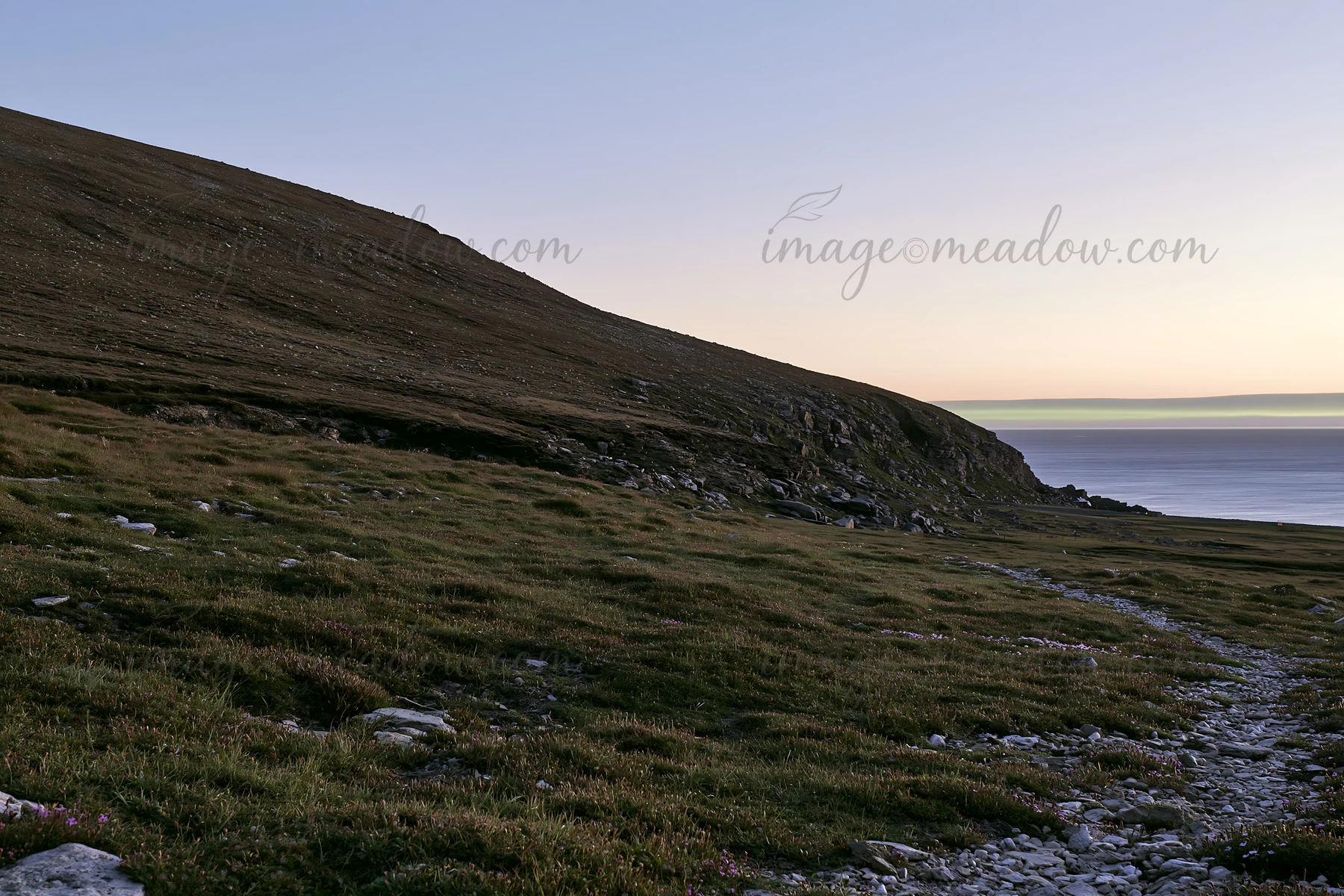 A rocky shoreline extends along the coast, surrounded by grassy meadows. The low sun casts a warm glow, marking the transition from day to night. The landscape