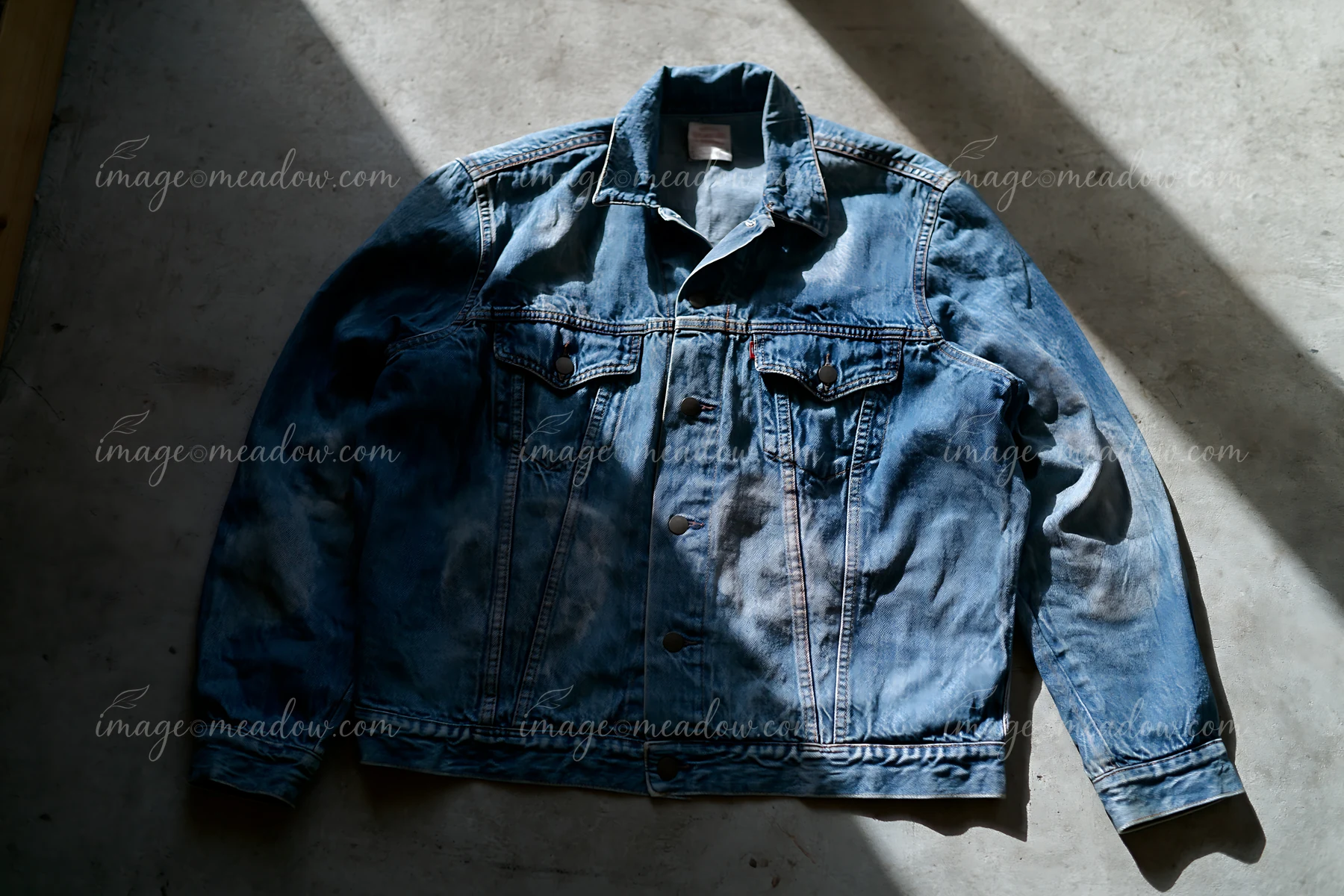 Blue denim jacket with dried watermarks lies on a concrete floor in strong diagonal sunlight.