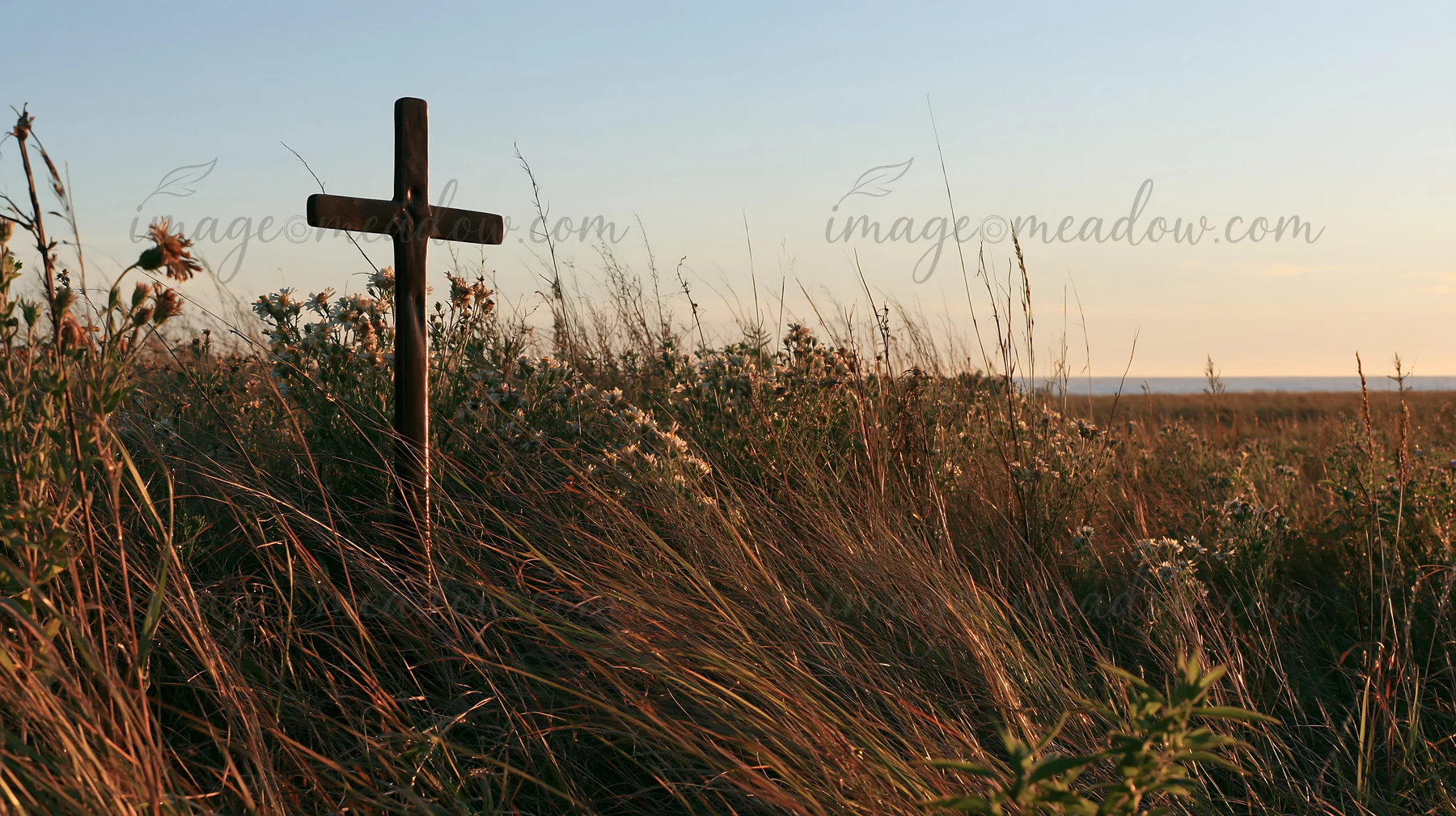 A bronze Christian cross stands tall in a meadow filled with tall grass and wildflowers during the Christmas season. The sun sets in the background, casting war