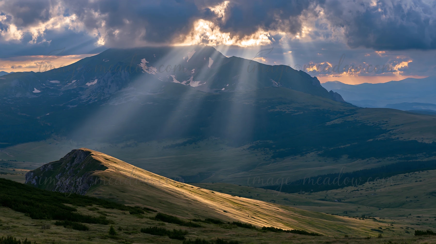 Light rays shine down on a meadow in the Bucegi Mountains. The scene shows rolling hills and distant peaks under a cloudy sky. The time of day appears to be lat