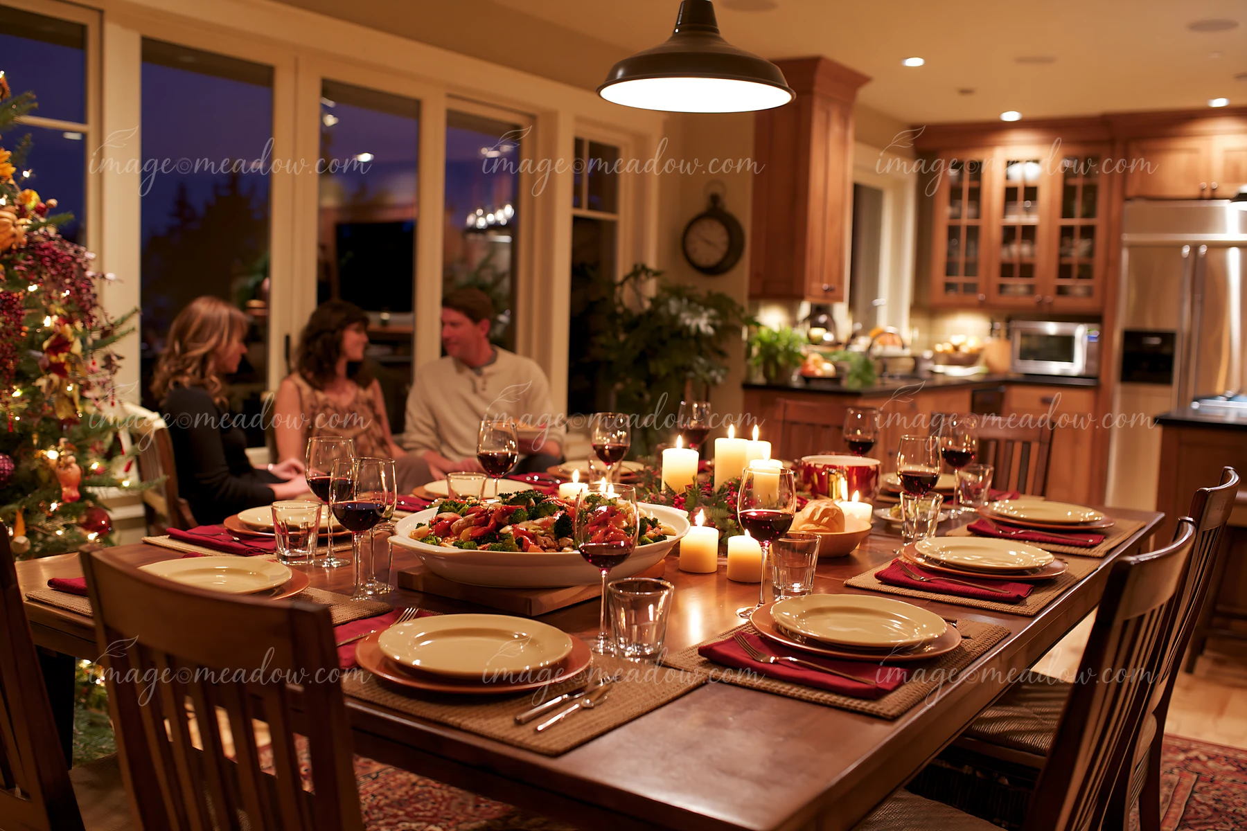 Candlelit holiday dining table set with food and wine glasses, while three people sit near a decorated Christmas tree.