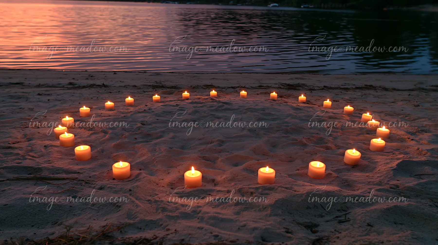Candles arranged in a circle are placed on the sand by a lake during twilight.