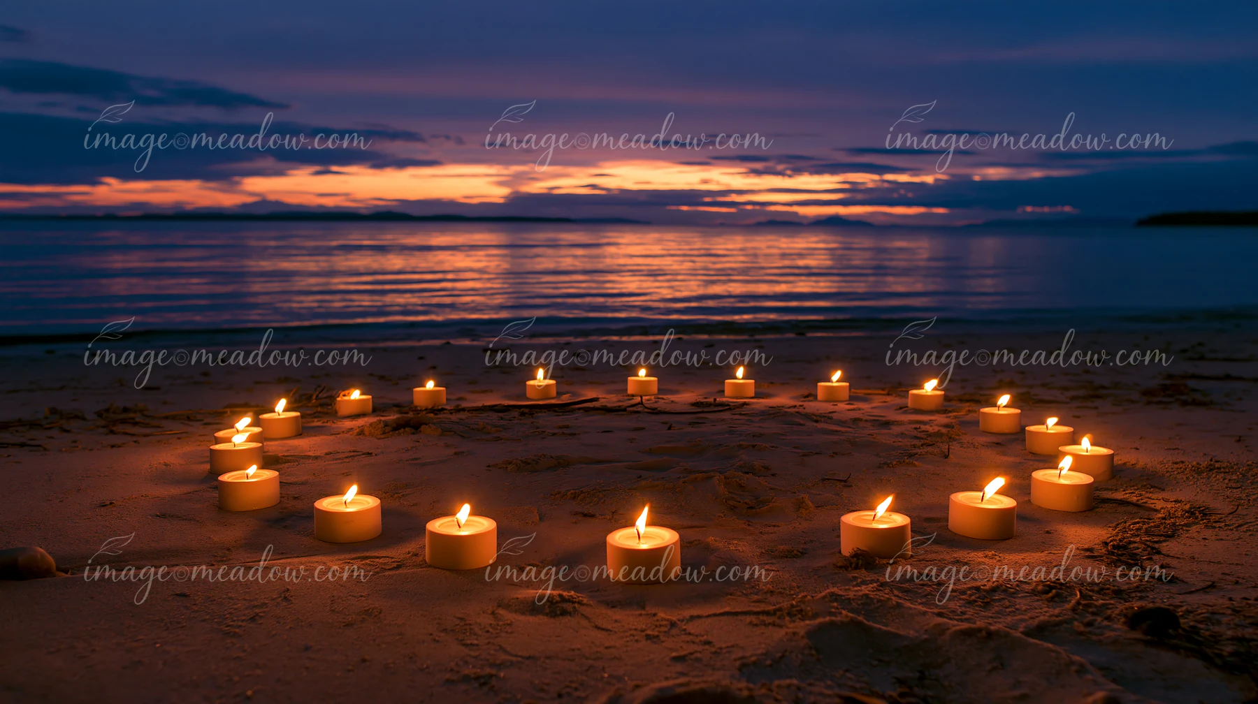 Candles arranged in a circle on a sandy beach illuminate the scene as the sun sets over the water.