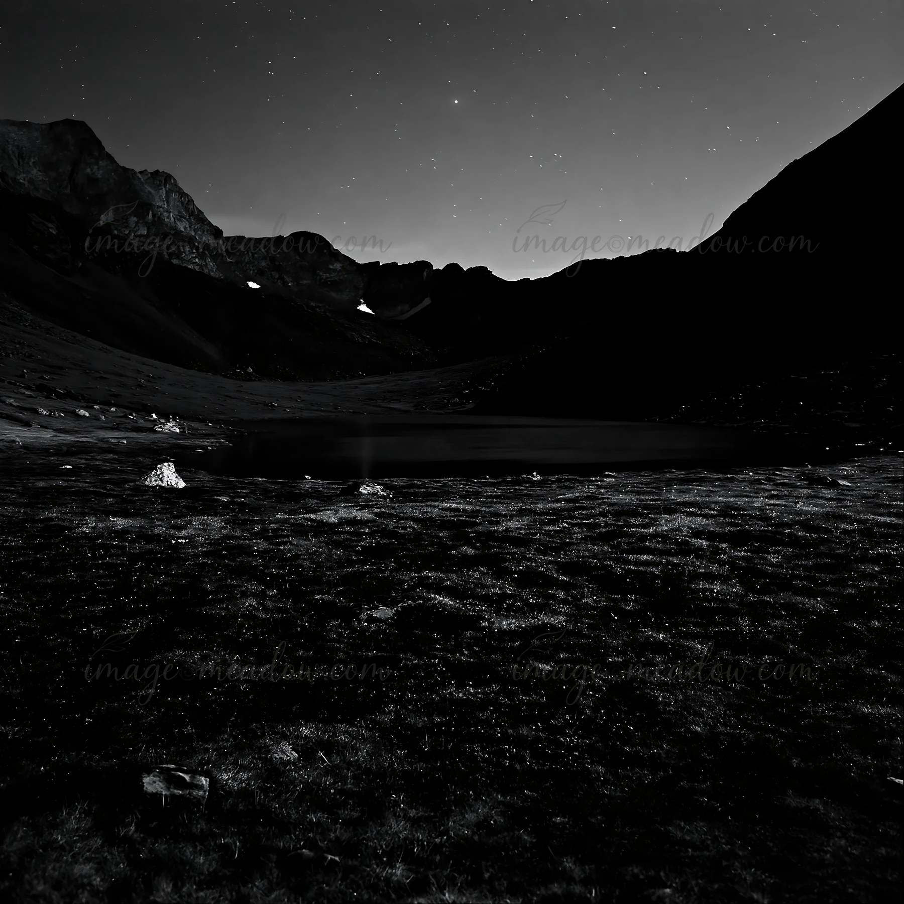 A dark landscape features a lake surrounded by mountains and rocks under a starry sky. The scene captures a quiet setting at nighttime, highlighting the natural