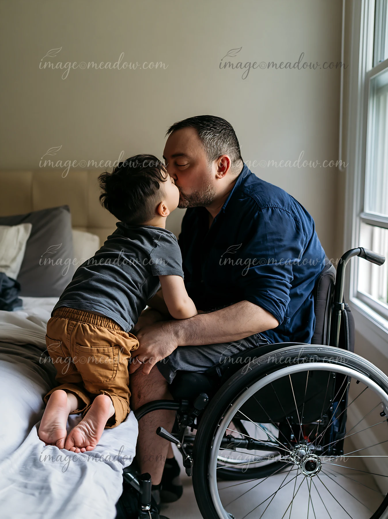 Child kisses a seated father in a wheelchair beside a bed in a softly lit bedroom.