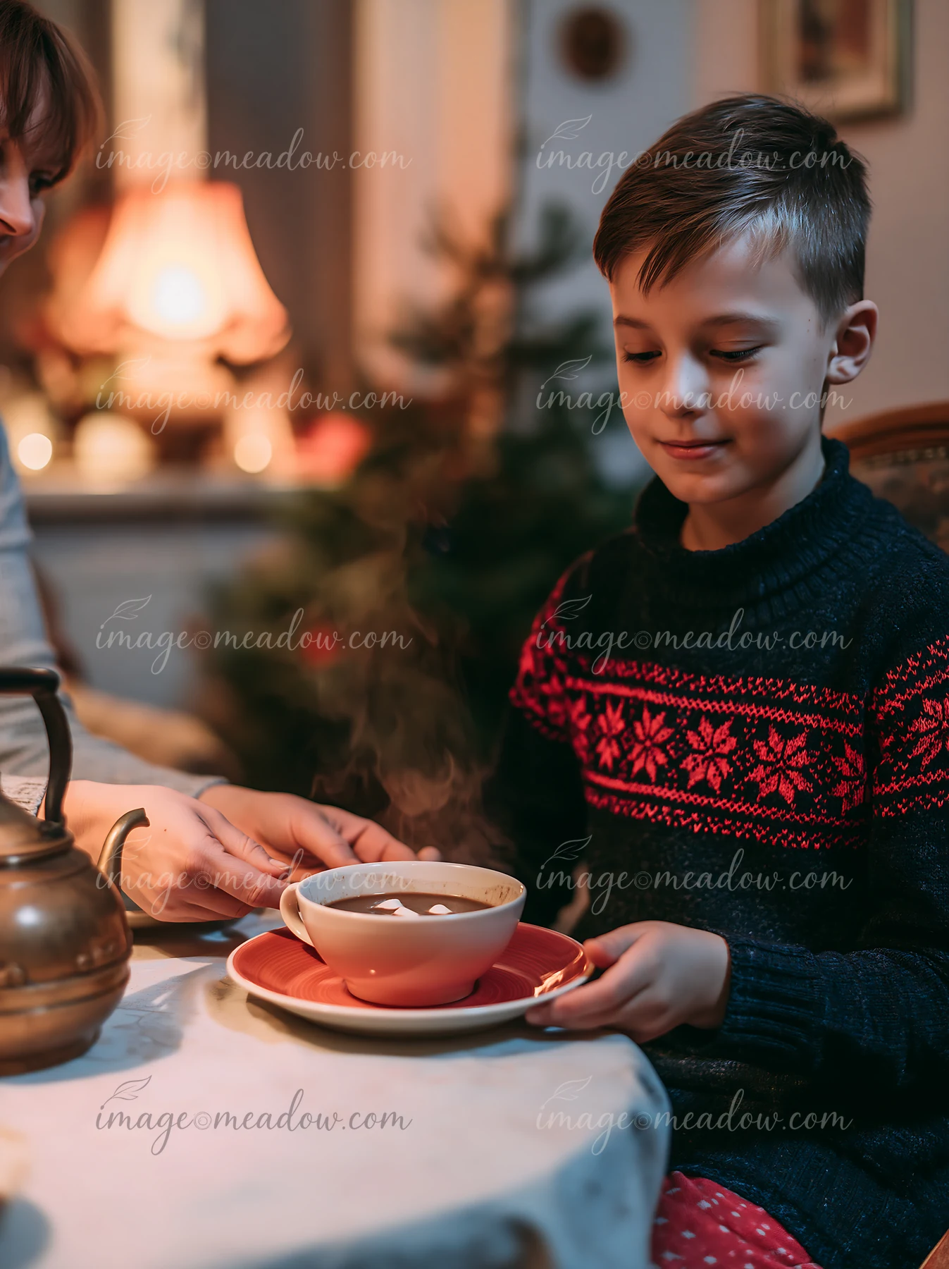 Child sits beside a Christmas tree while an adult serves steaming cocoa with marshmallows at a table.