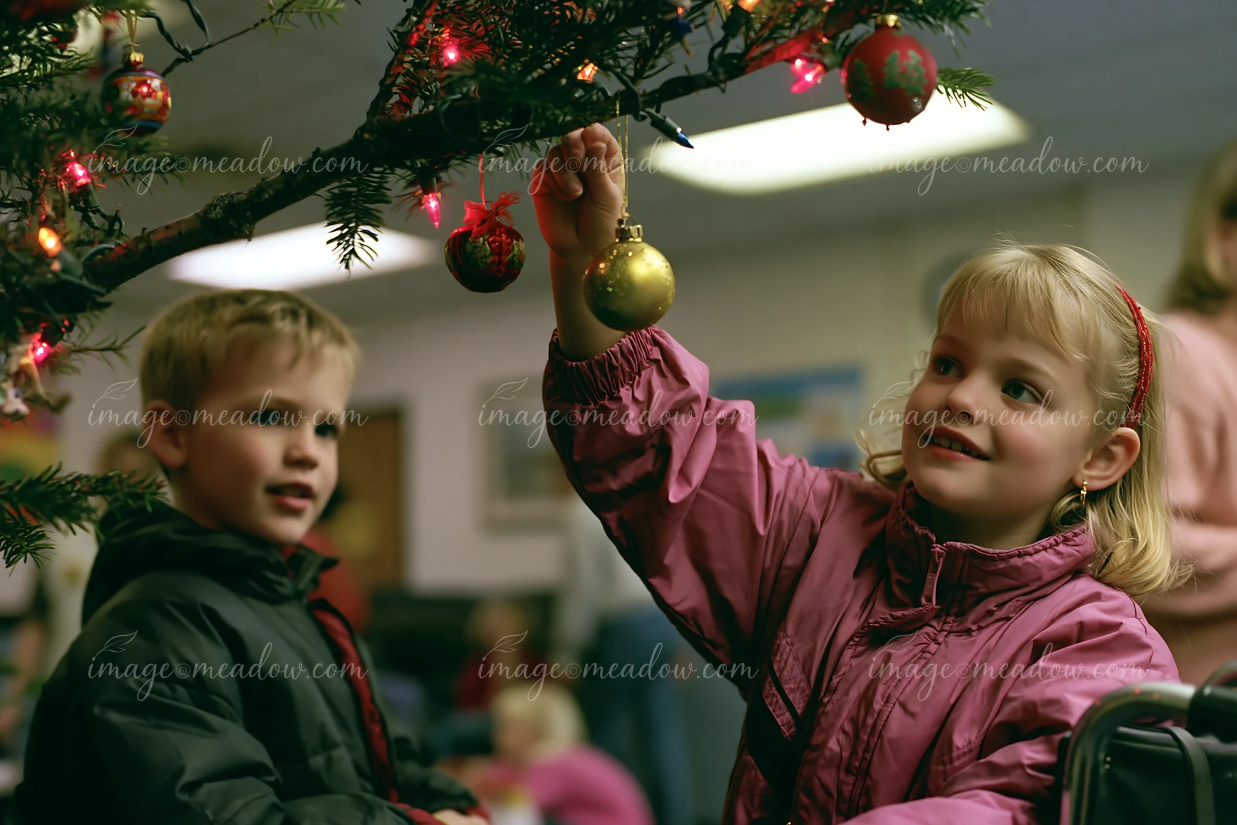 Children hang colorful ornaments and lights on Christmas tree branches indoors.