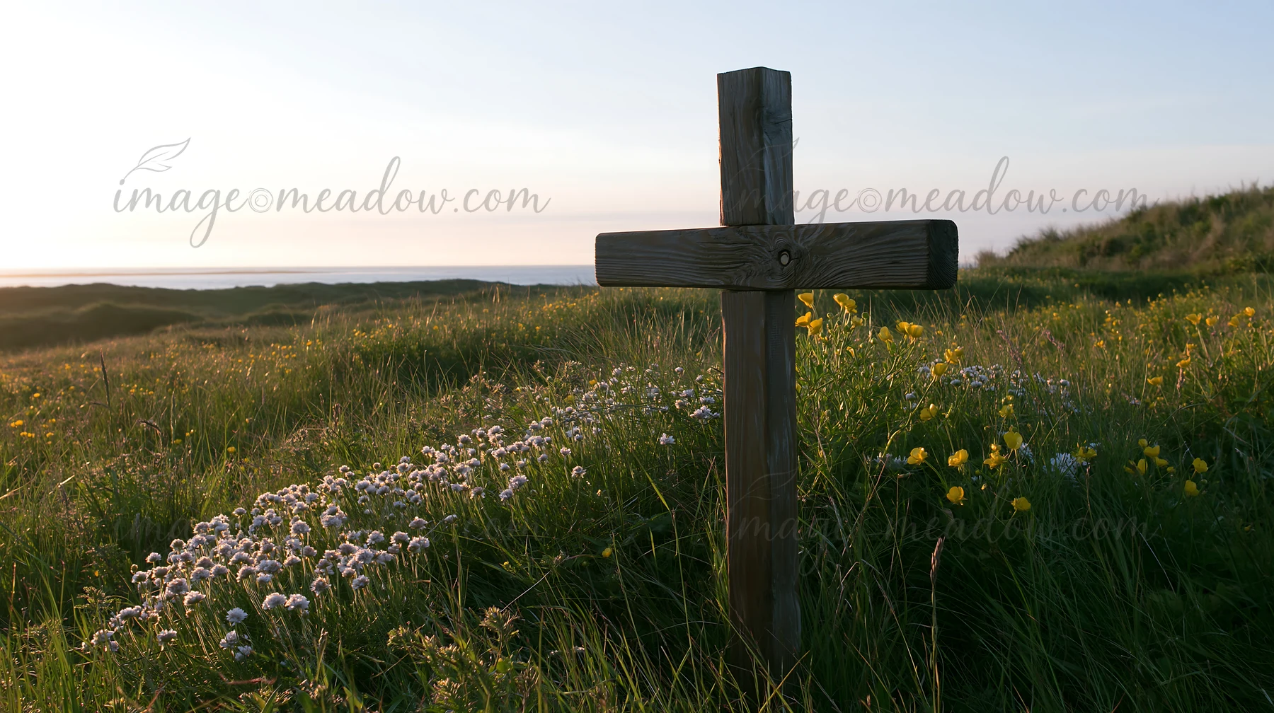 A wooden cross stands in a meadow filled with flowers as the sun rises. The scene captures the essence of dawn with soft light illuminating the grass and blooms