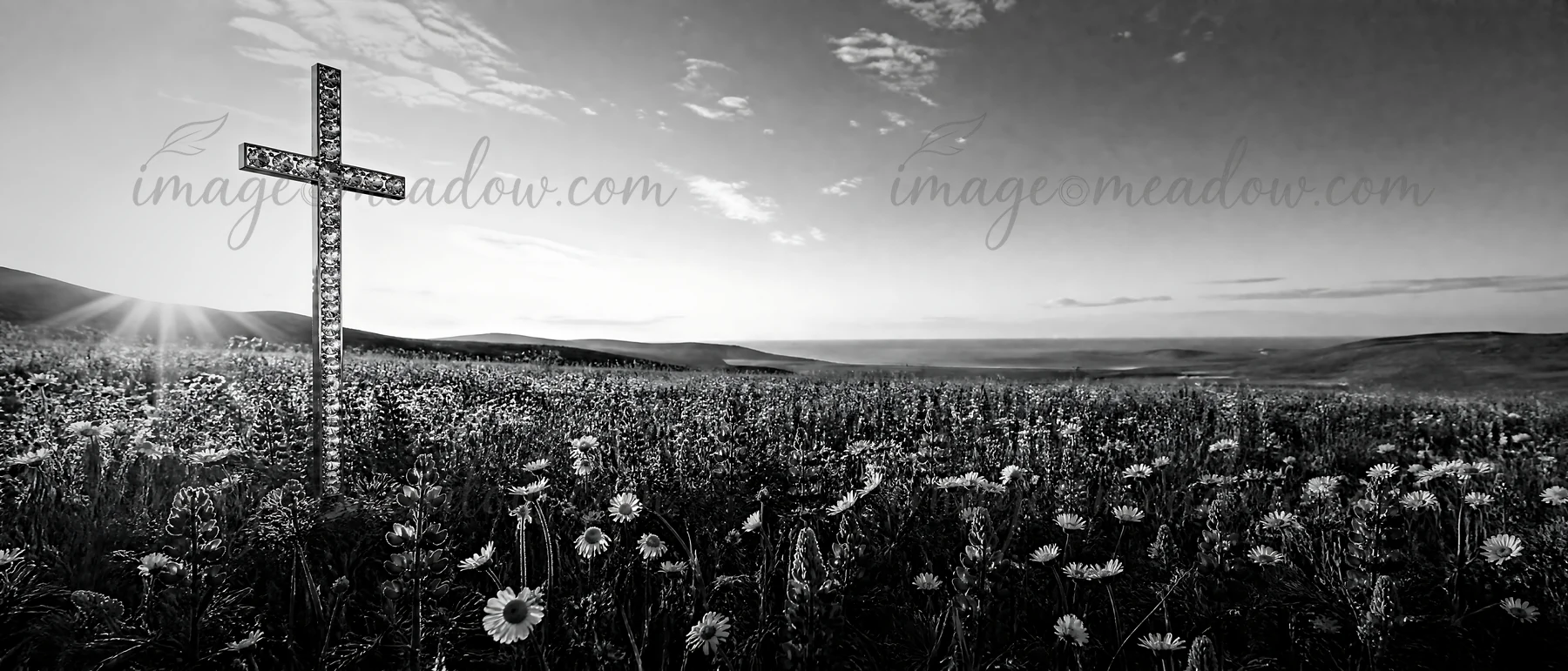 A cross stands in a meadow filled with flowers as light rays shine through the sky. The scene captures a moment of daylight in an open field surrounded by hills