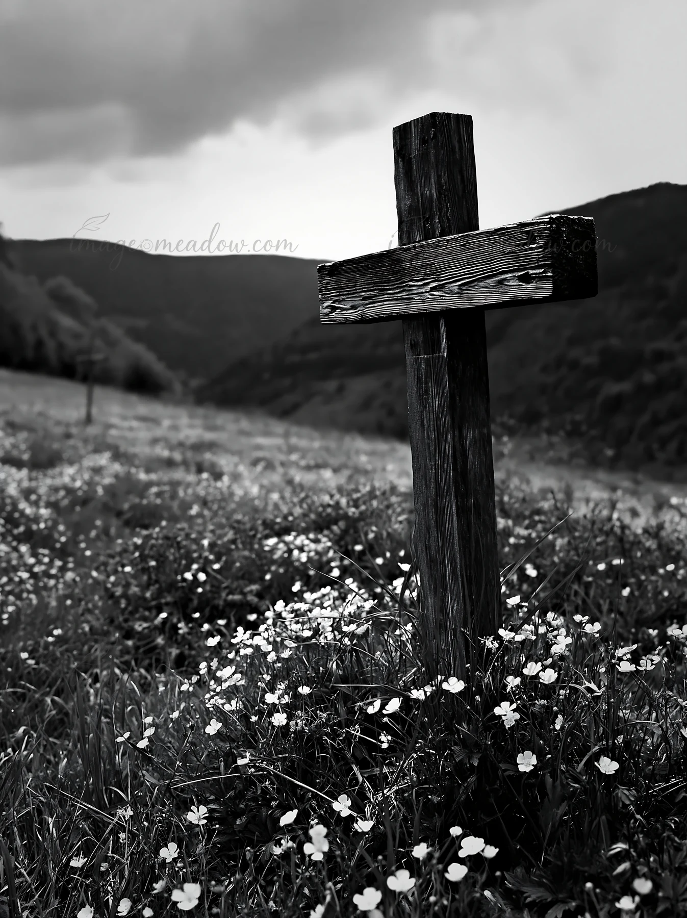 A wooden Christian cross stands in a meadow filled with flowers. The landscape shows rolling hills in the background under cloudy skies. This scene captures a m