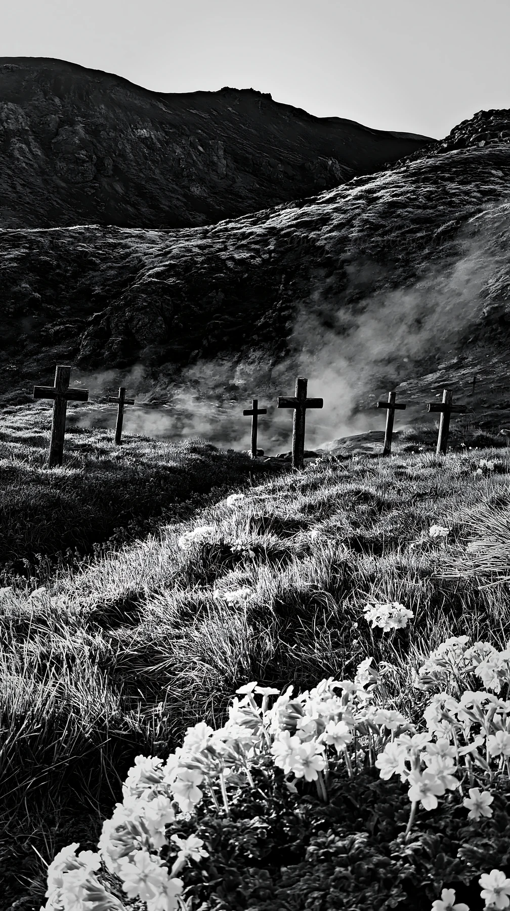 Christian crosses stand in a meadow near a geothermal spring, surrounded by green grass and flowers. The setting features smoke rising from the spring, with mou
