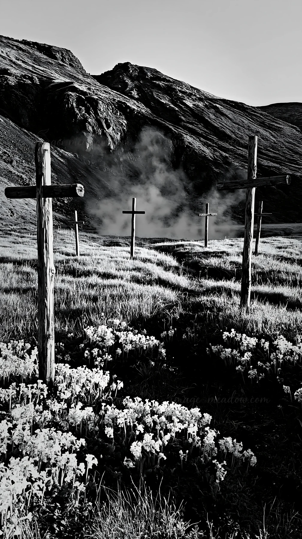 A group of wooden crosses stands in a meadow near a geothermal spring. The landscape features grassy terrain and a backdrop of mountains. Steam rises from the s