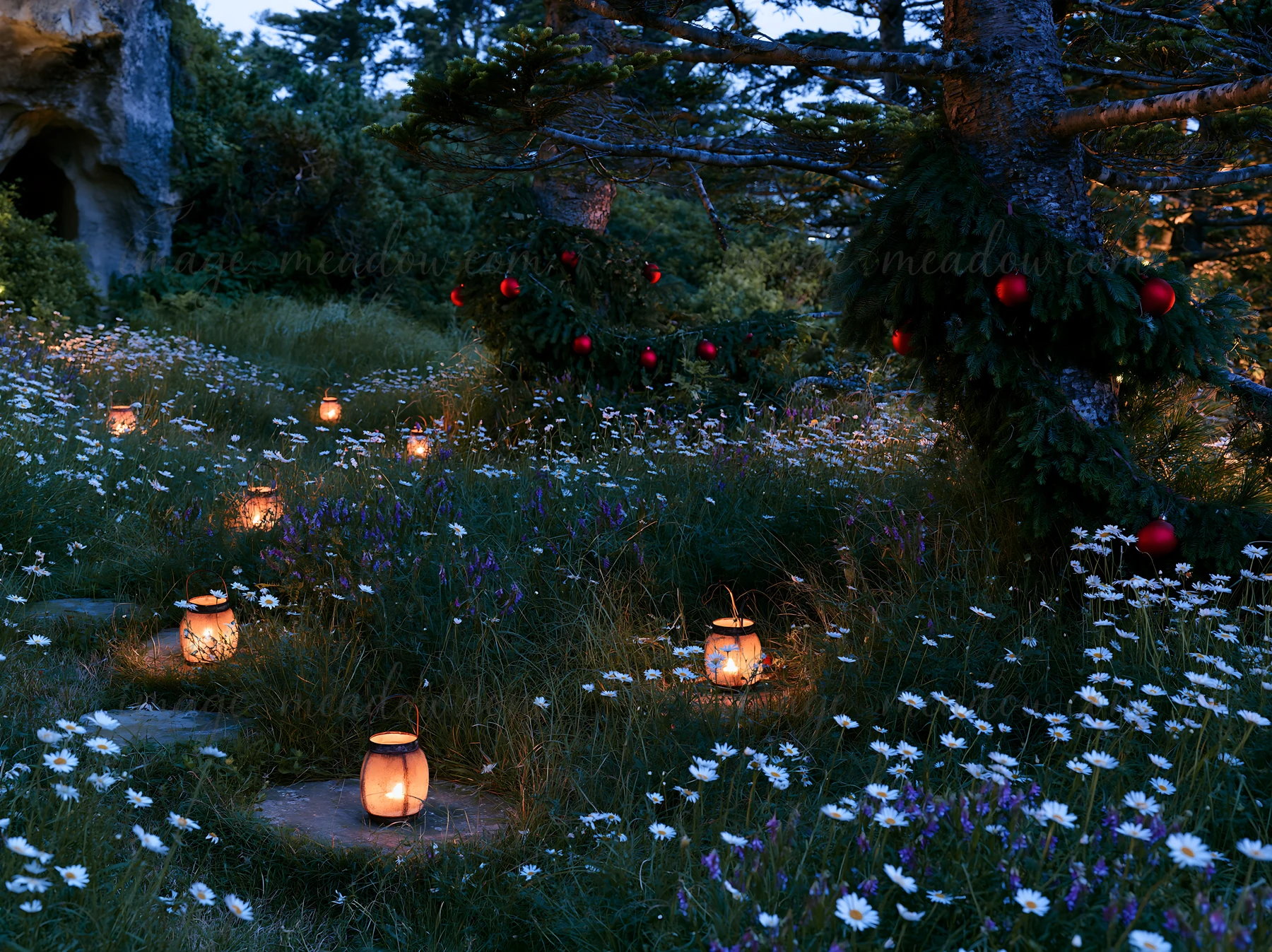 Lanterns glow softly in a meadow surrounded by trees and flowers at dusk. A cave is visible nearby, adding to the atmosphere of the setting. The space is decora