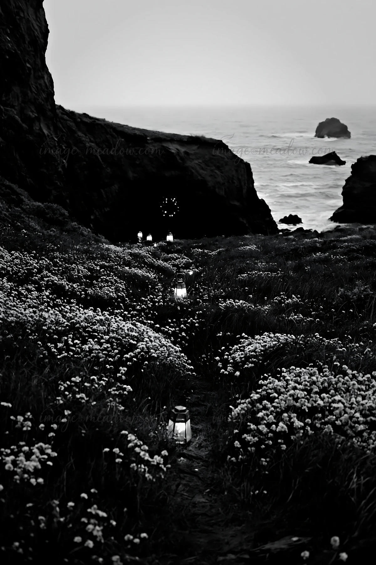 A pathway lined with lanterns leads to a cave in a meadow close to the ocean. The scene is dusk with low light, highlighting the soft glow of the lights among t