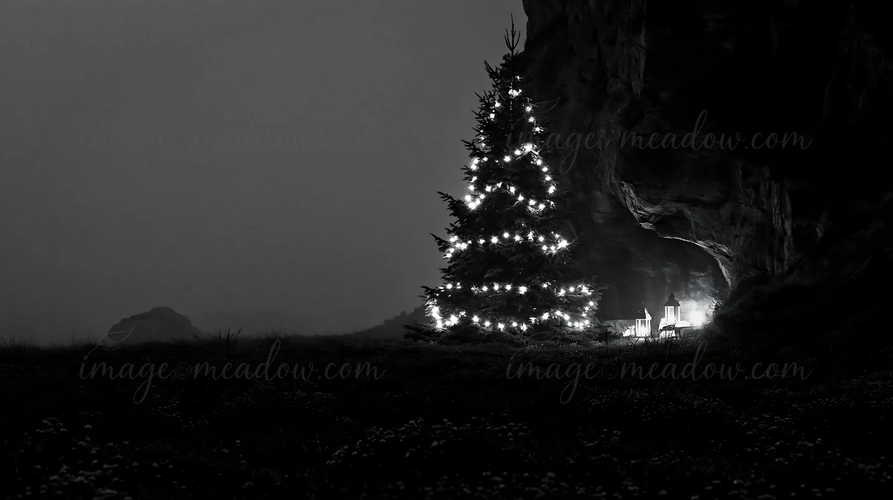A Christmas tree is illuminated with lights, standing in a meadow next to a cave during dusk. The scene captures a tranquil moment in nature with soft lighting