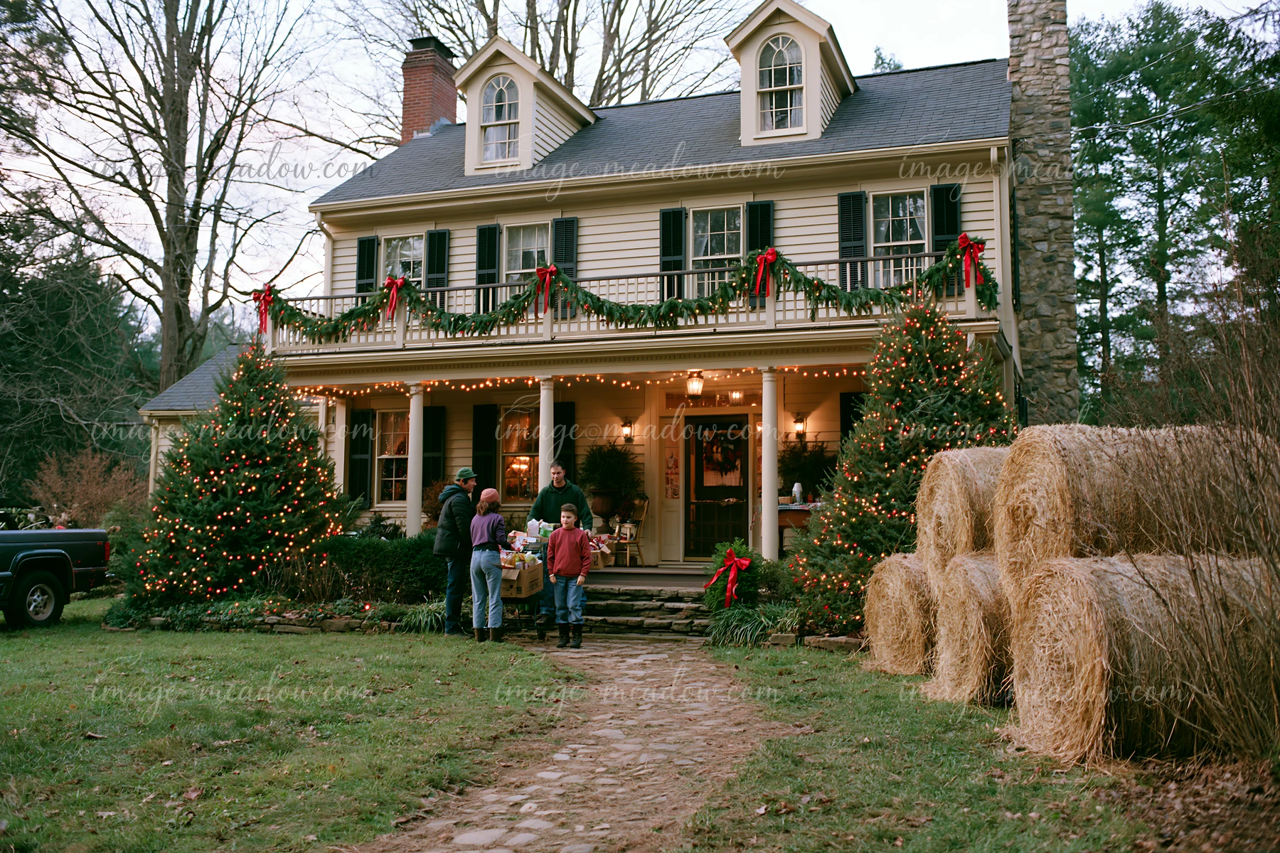 Country house decorated with pine garlands, lit Christmas trees, hay bales, and four people outside.