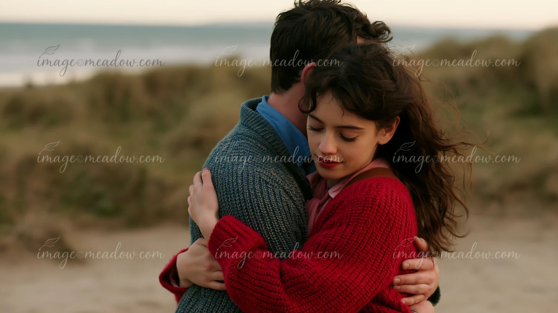 Couple embracing closely on a sandy beach with dunes and sea in the background.