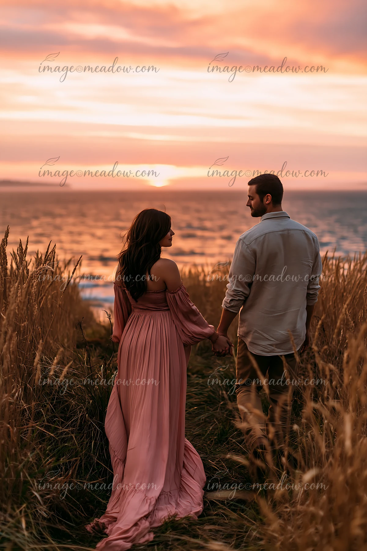 Couple holding hands in tall coastal grass beside the ocean as the sun sets.