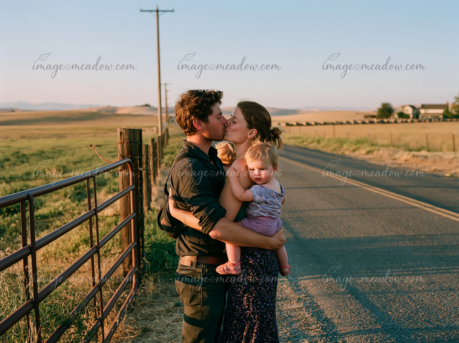 Couple kisses beside a country road while holding a baby, with farmland and a fence behind them.