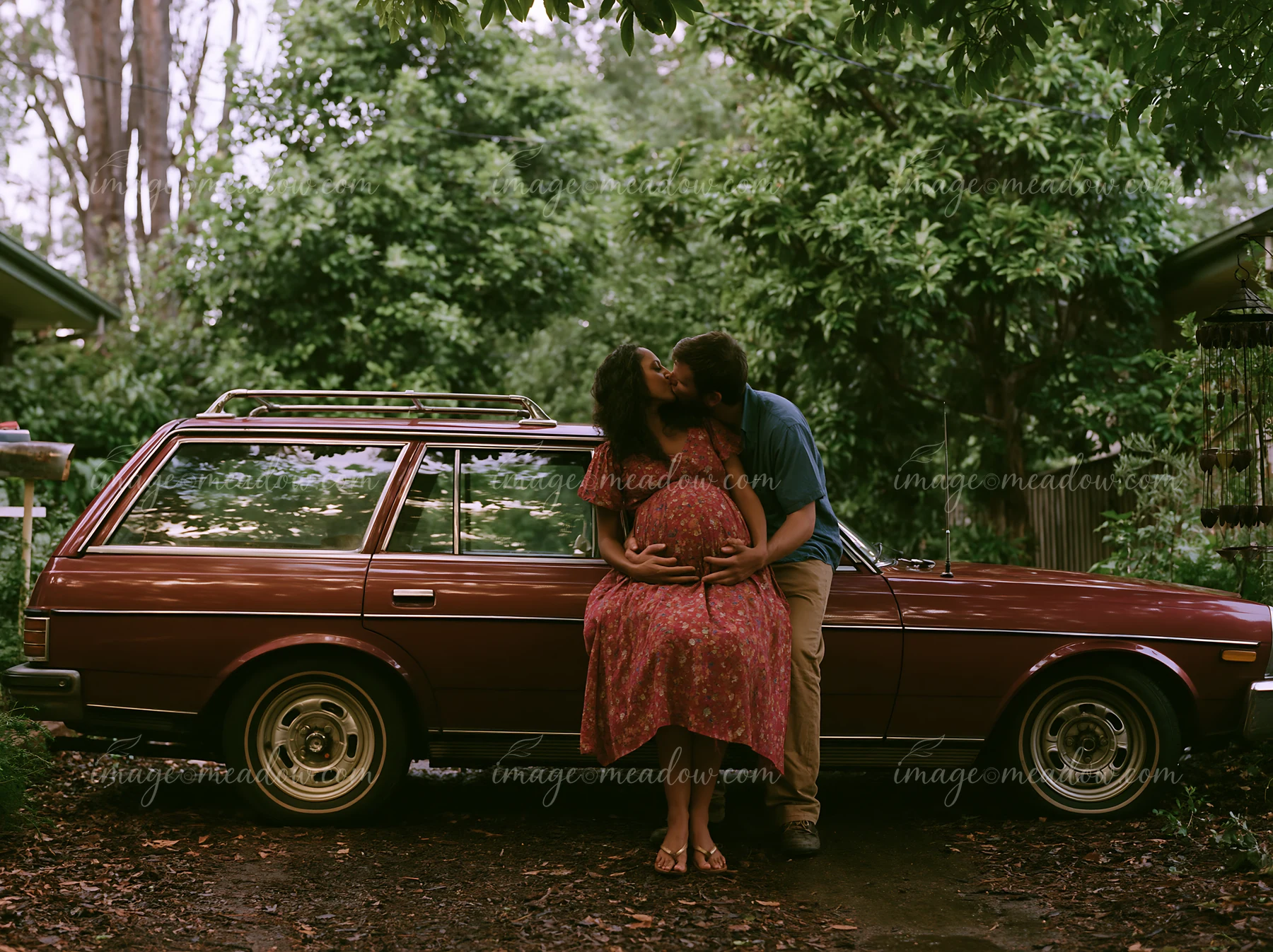 Couple kisses beside a vintage station wagon in a leafy wooded setting.