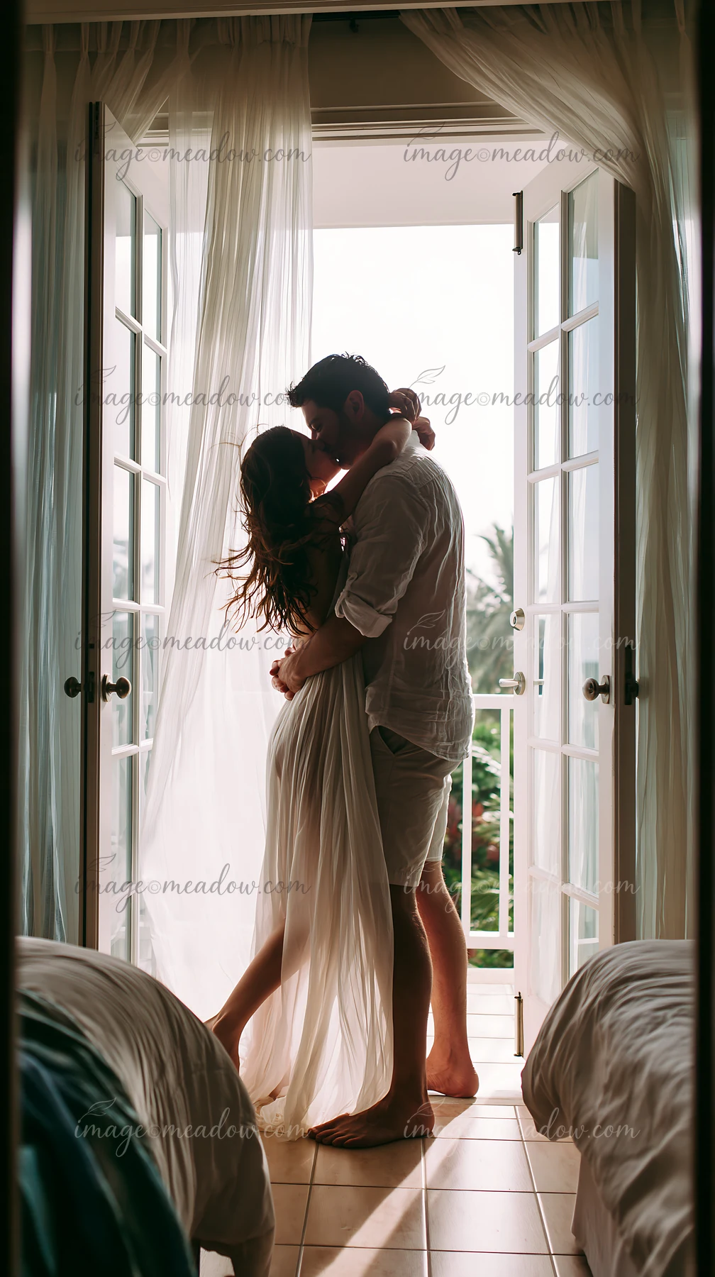 Couple kisses beside open French doors in a bright bedroom with sheer curtains.