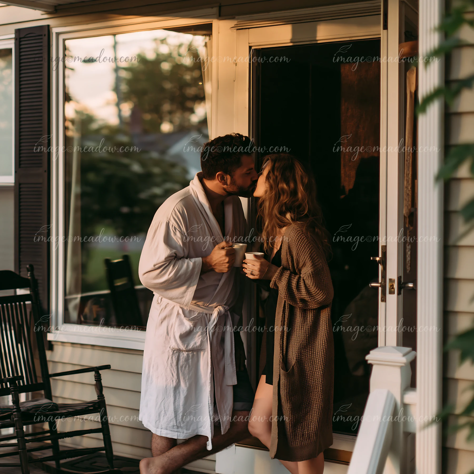 Couple kisses while holding coffee cups beside a sunlit open doorway at home.