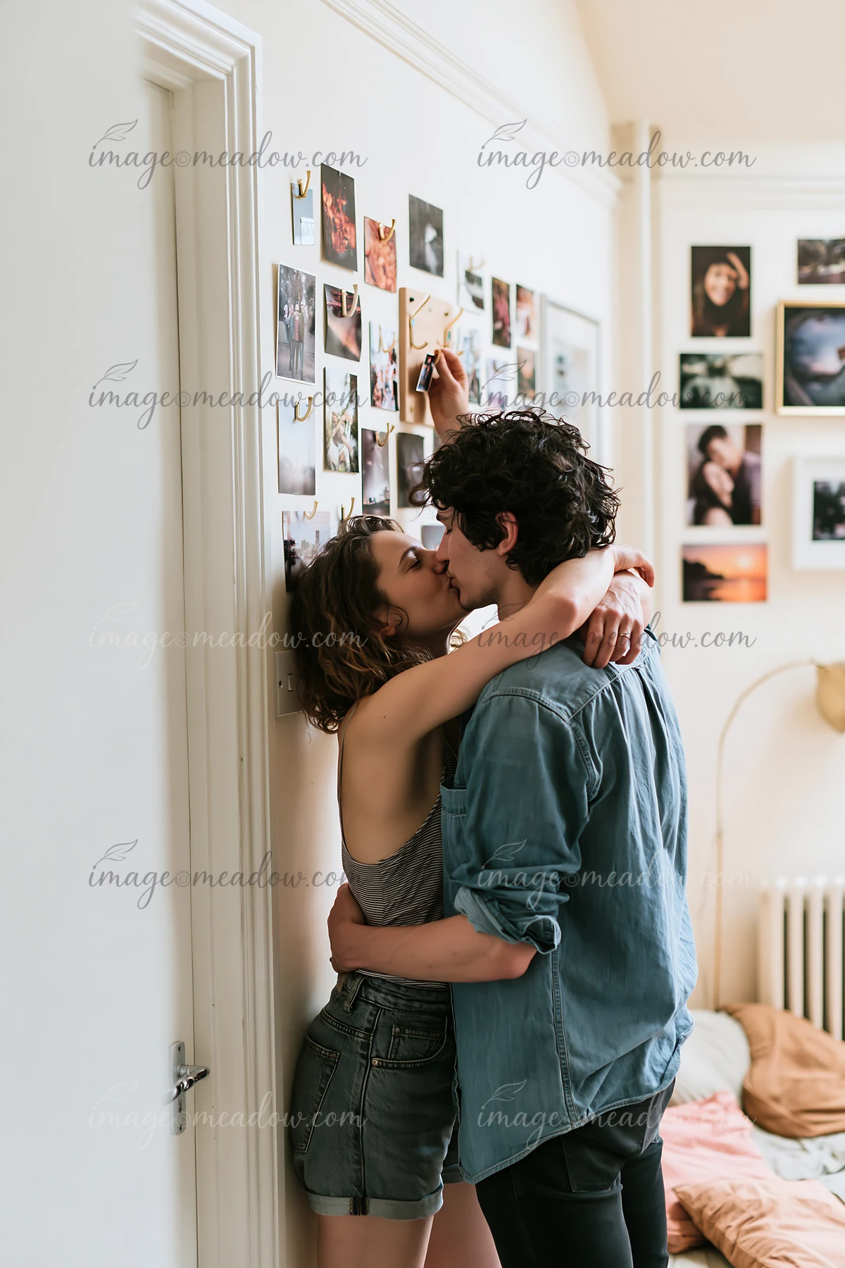 Couple kisses while one partner decorates a photo wall in a bright bedroom.