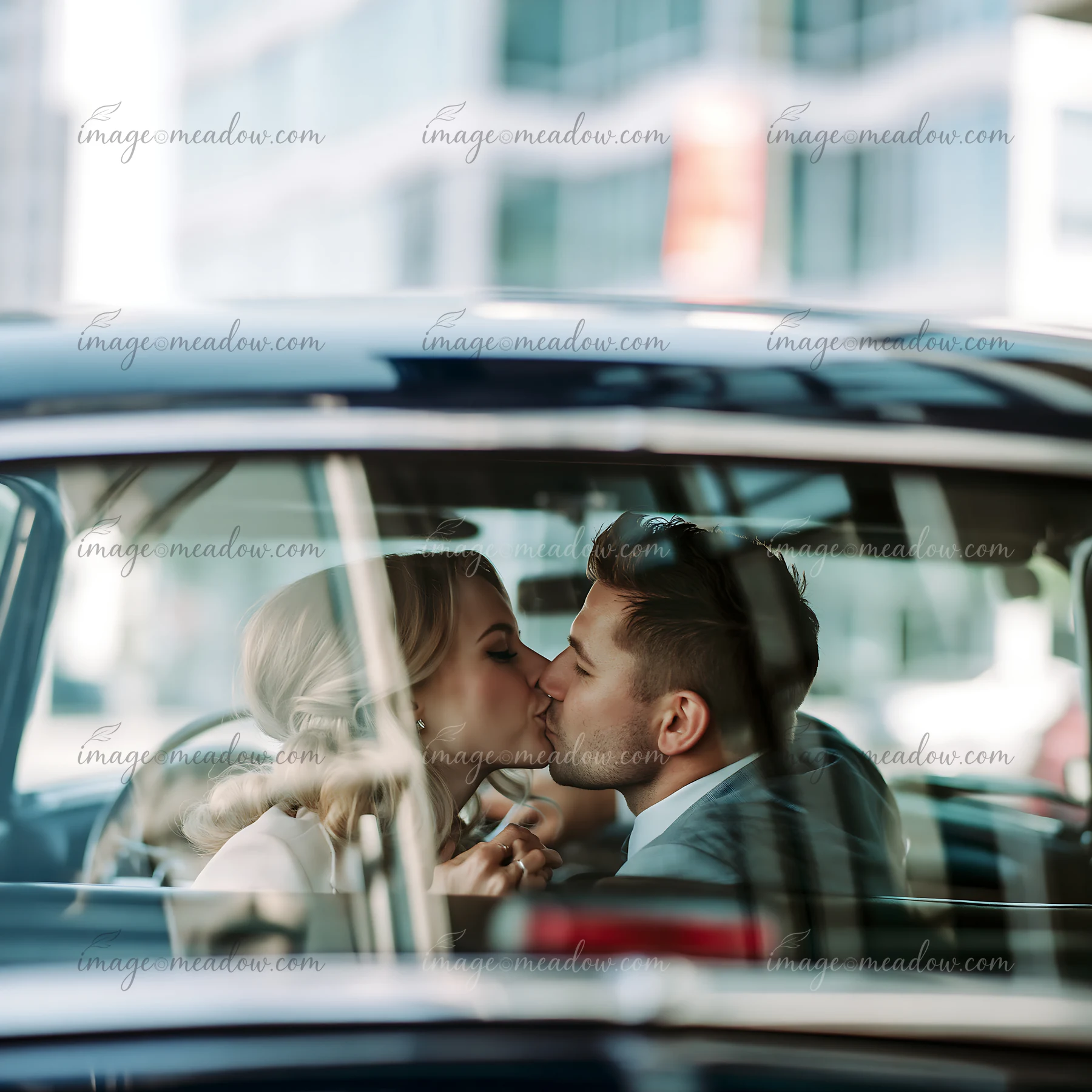 Couple kissing inside a vintage car, seen through the windshield with blurred buildings in the background.
