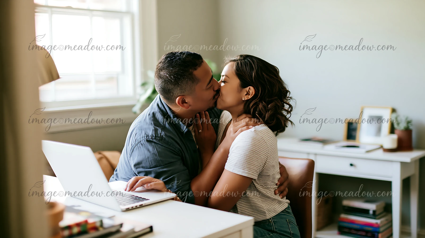 Couple shares a tender kiss beside an open laptop in a bright home office.