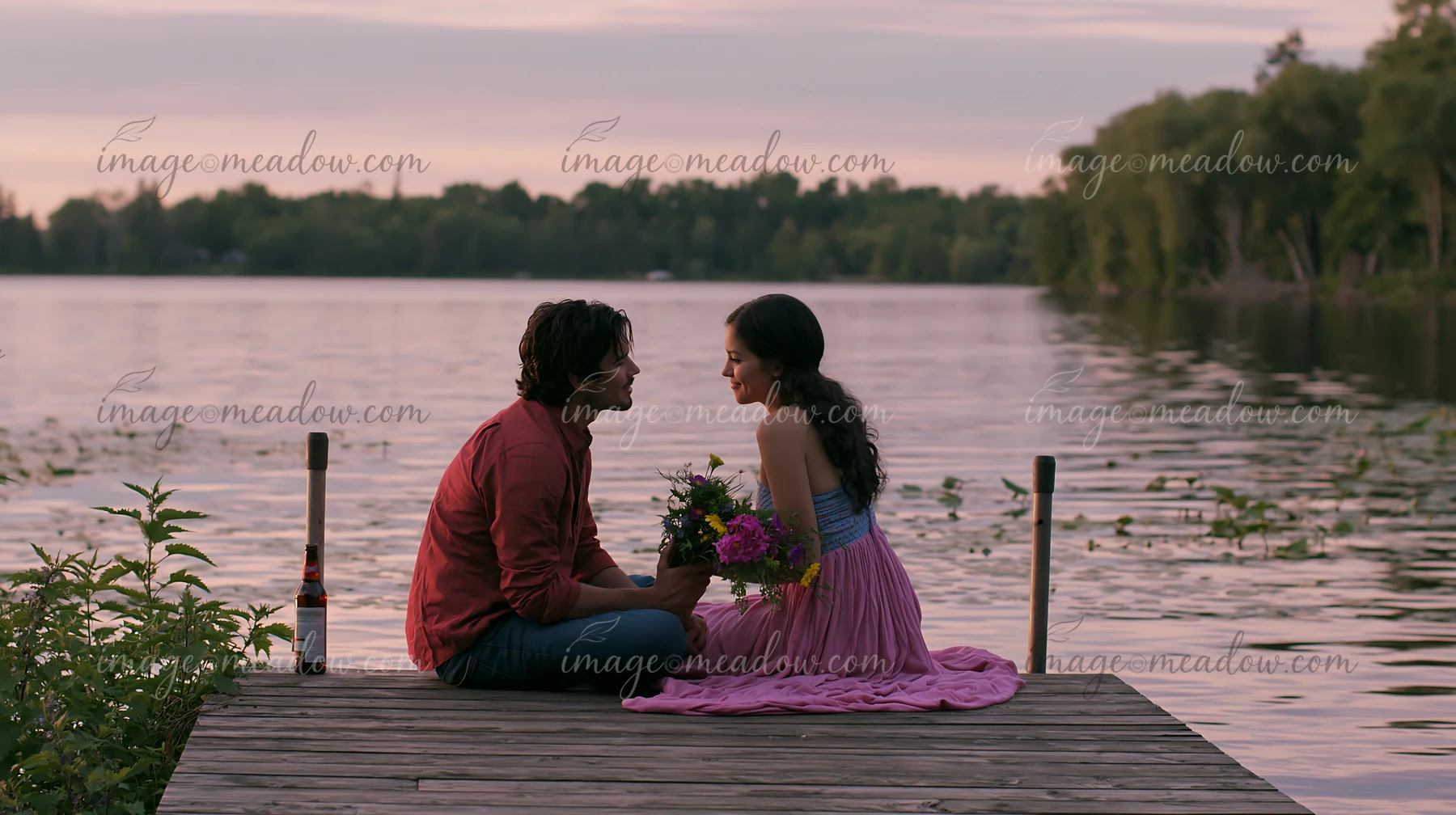 Couple sits together on a lakeside dock at sunset with flowers and a bottle nearby.