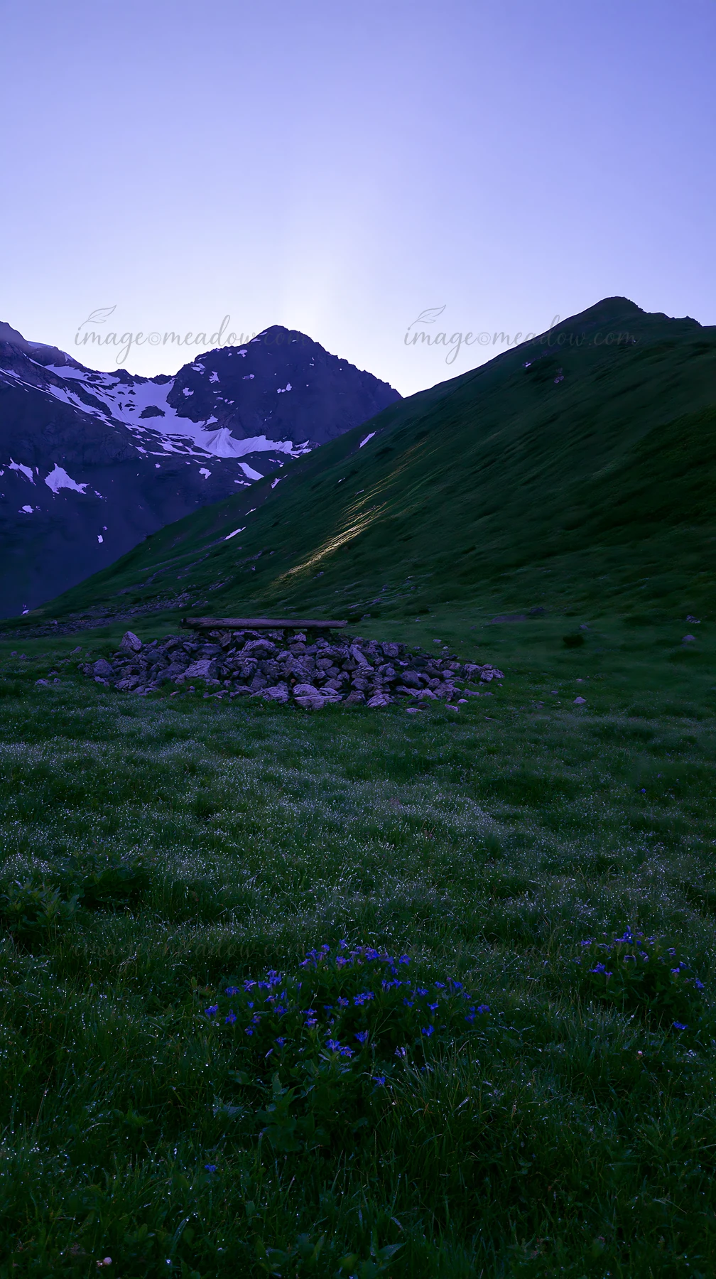 Crepuscular rays illuminate a lush green meadow surrounded by mountains. Snow remains on the peaks as the sky transitions from day to night. Wildflowers add col