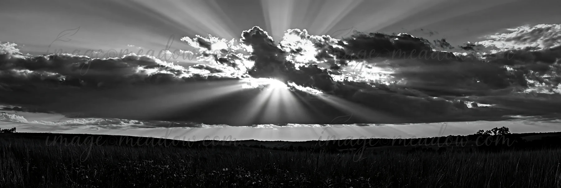 Sunlight breaks through clouds, casting rays across a vast meadow. The scene shows the sky filled with clouds while the ground is mostly bare, depicting a natur