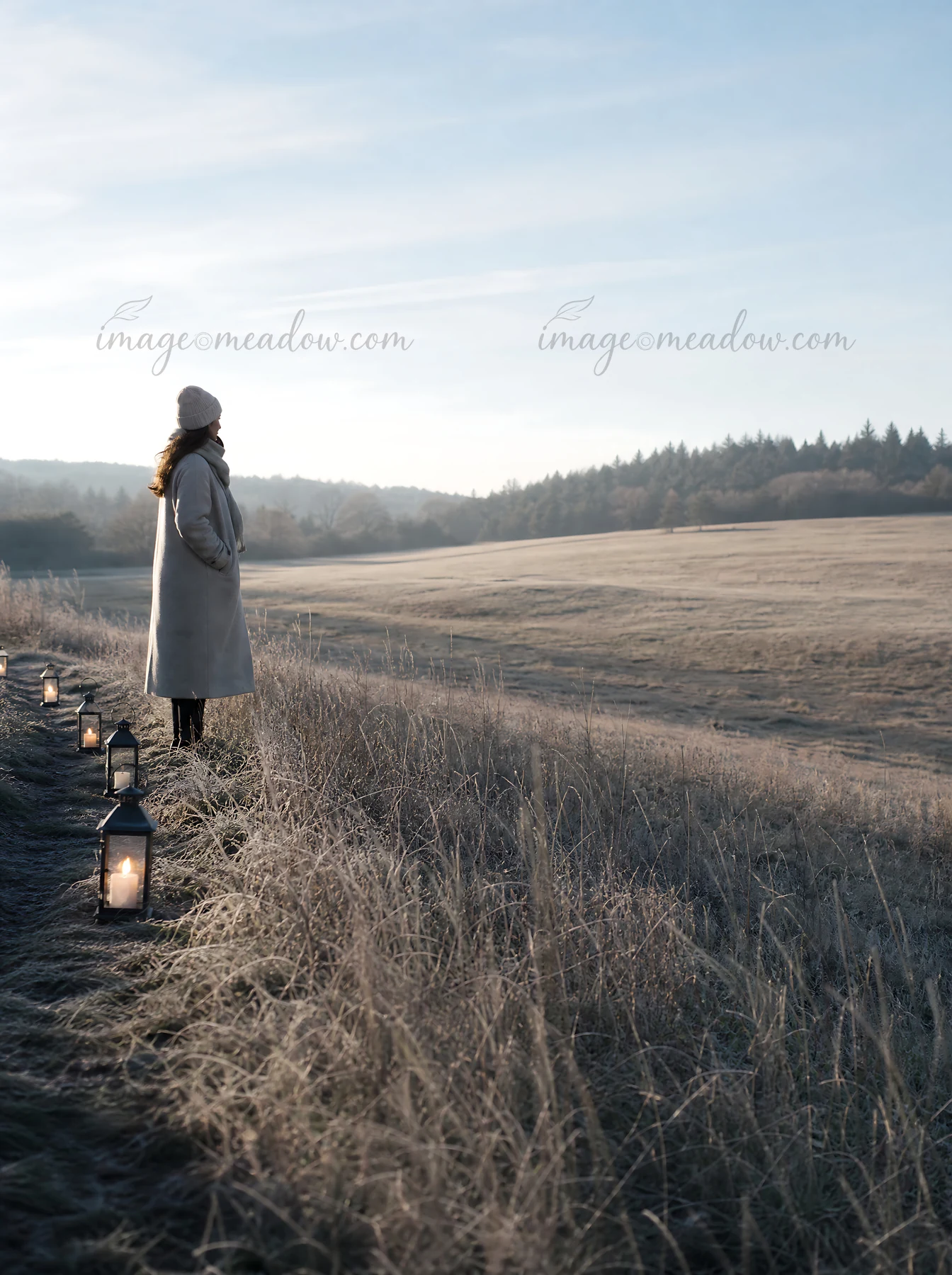 A person stands in a meadow observing the landscape on a clear day. Lanterns are placed along the path, creating a warm atmosphere against the natural backdrop