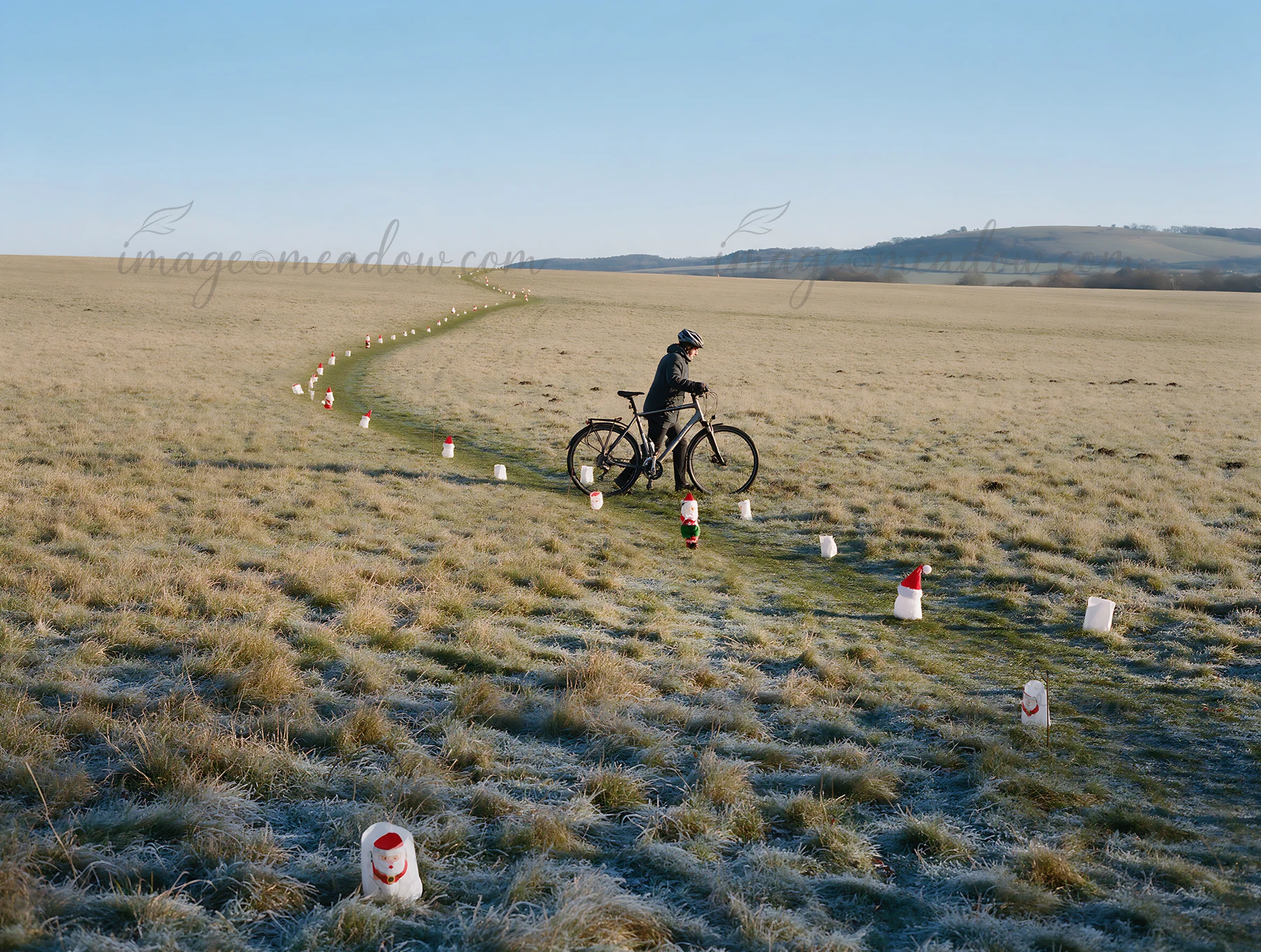 A cyclist rides through a meadow during daylight. The path is marked by colorful Christmas lanterns. The scene shows the frost-covered grass and distant hills.
