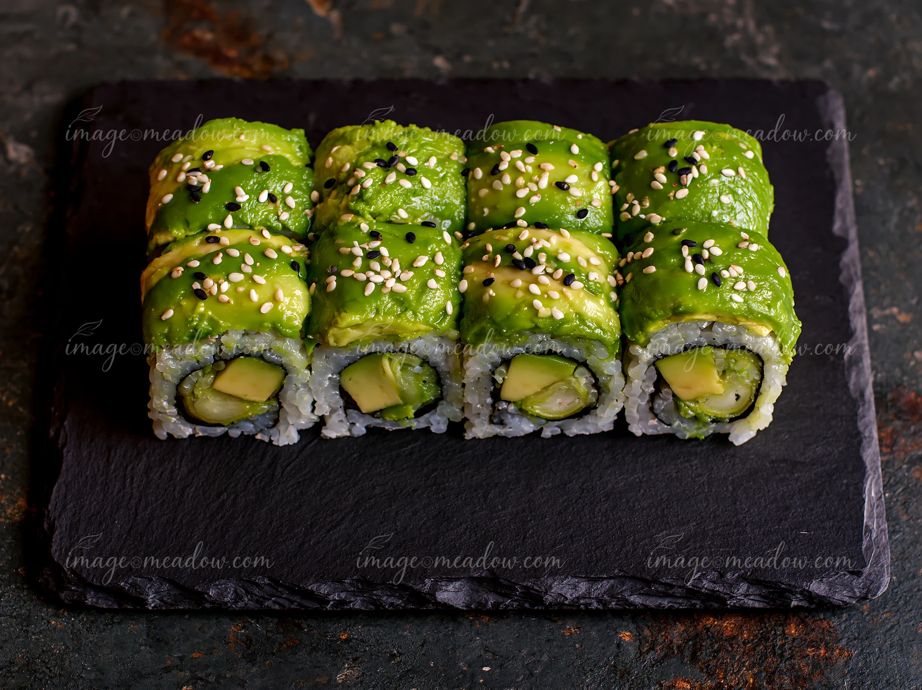 Dragon roll close-up features sushi rolls with avocado and cucumber, garnished with sesame seeds, served on a dark stone plate, ideal for food photography and s