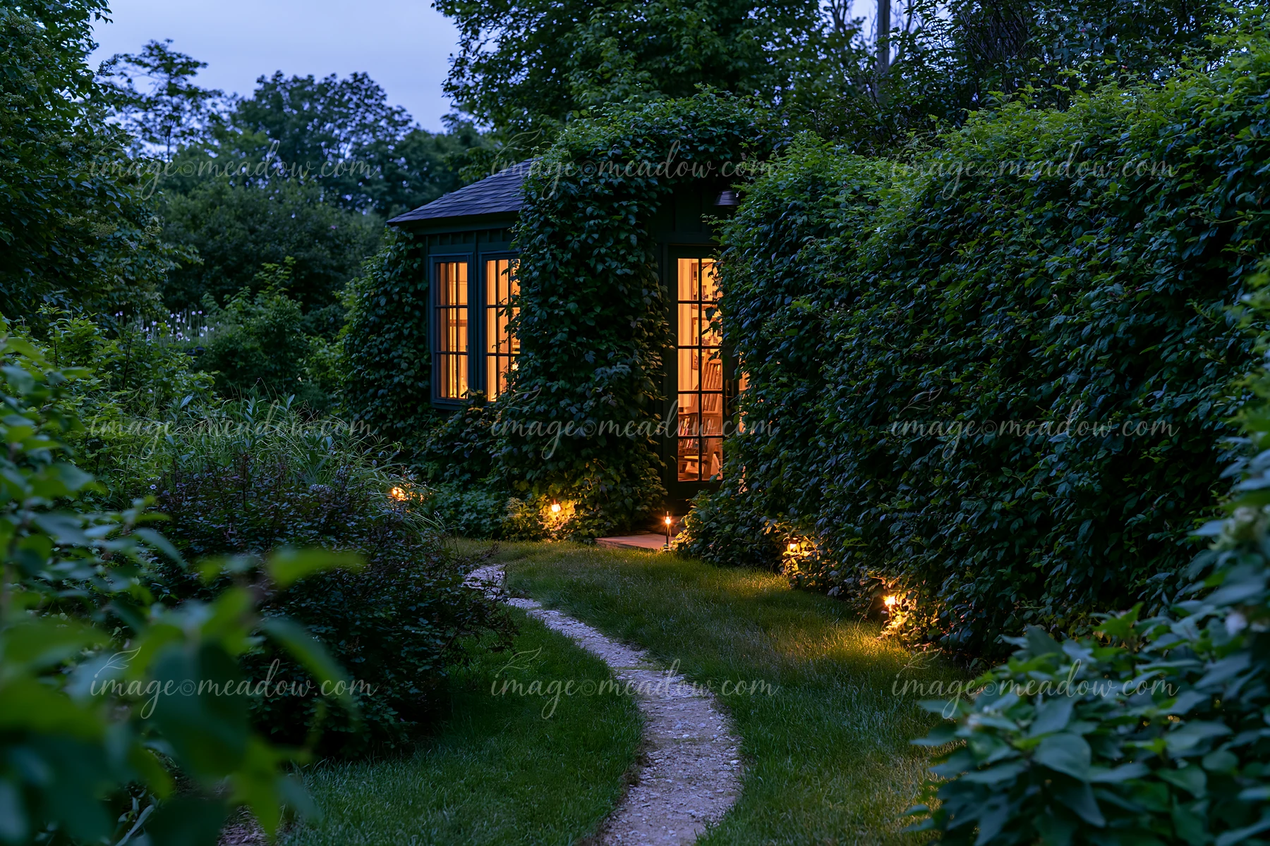 Empty garden studio glowing among dense greenery beside a warmly illuminated winding path at dusk.