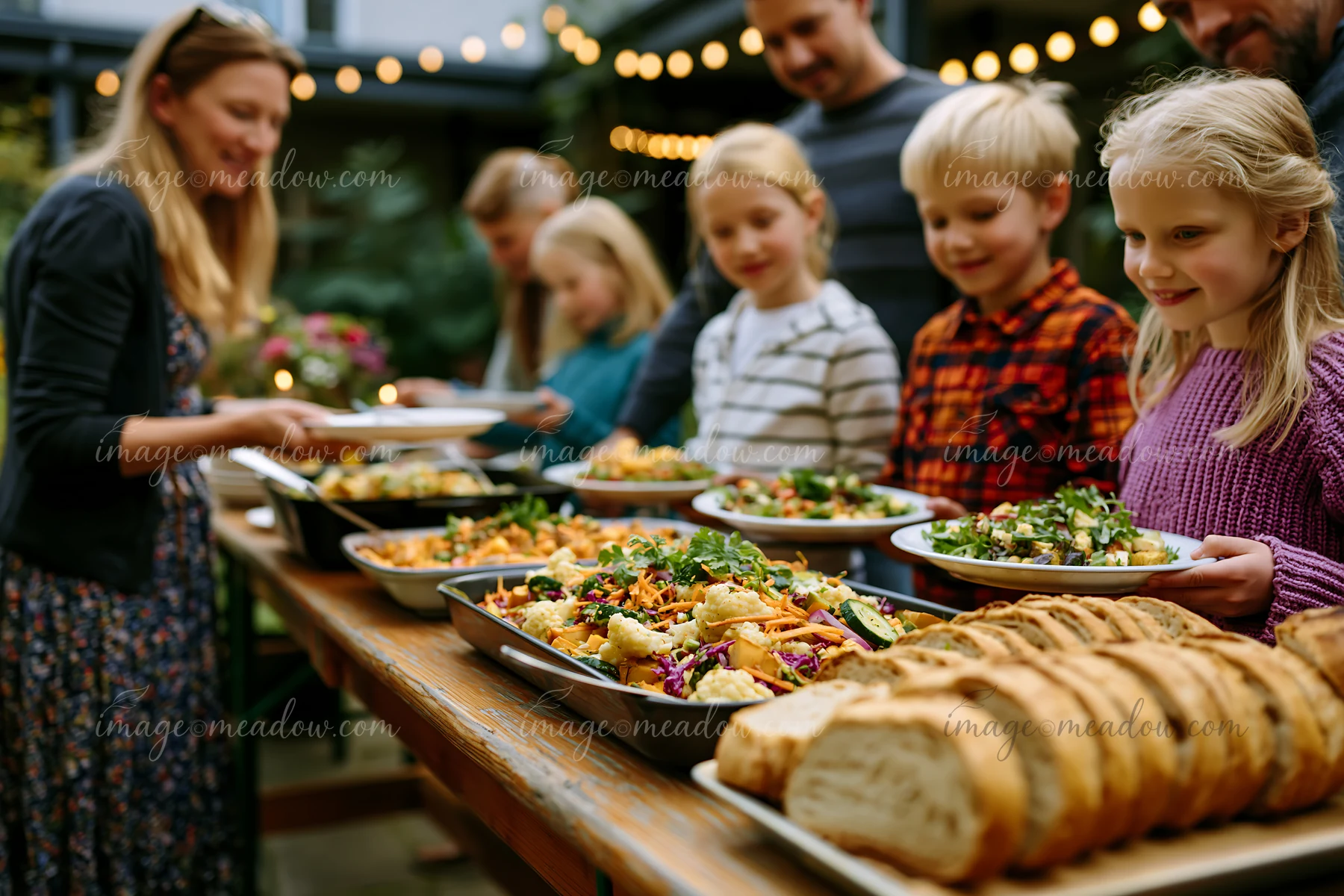 Families serve salads, vegetables, bread, and other dishes at an outdoor community potluck buffet.
