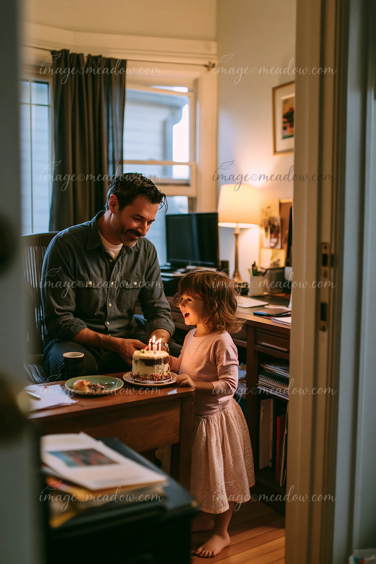 Father and young child celebrate around a lit birthday cake in a warmly lit home office.