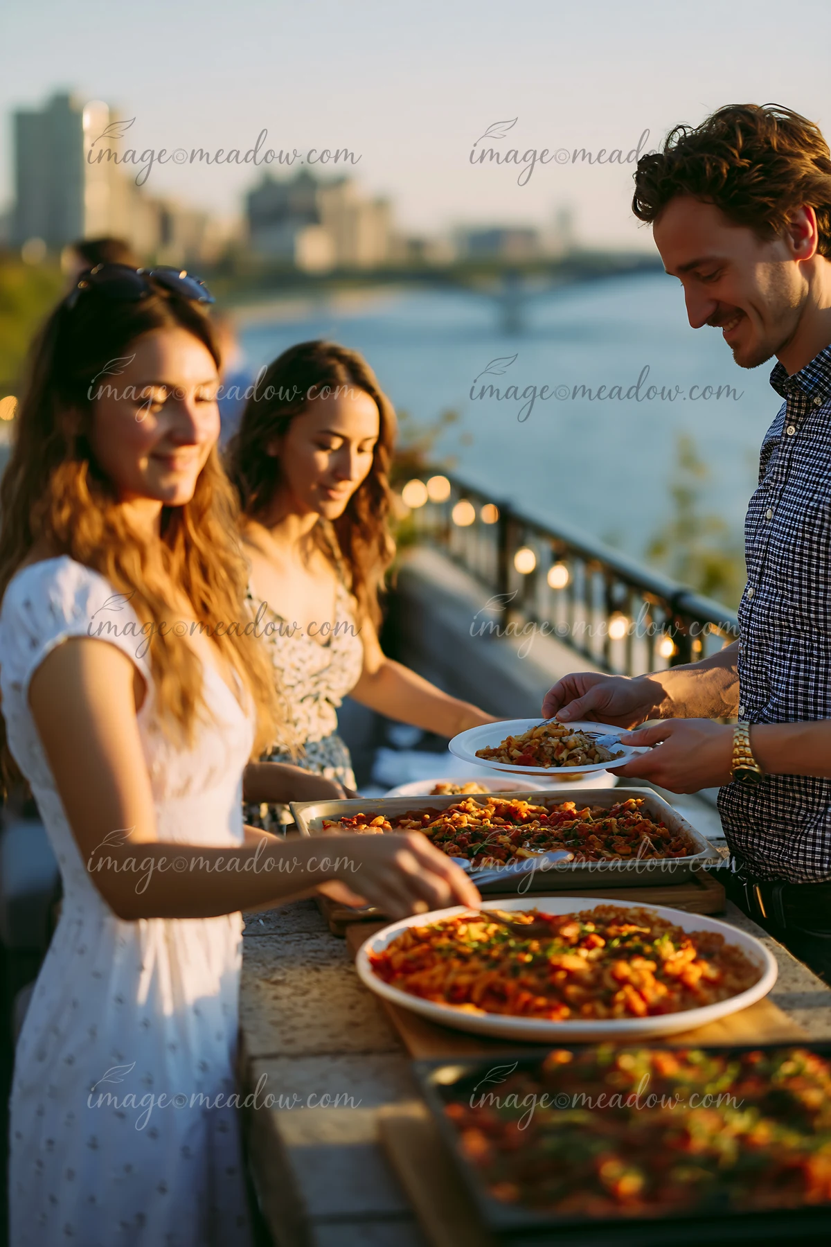 Friends serve themselves at a rooftop buffet dinner, enjoying a variety of dishes during sunset by the riverside.