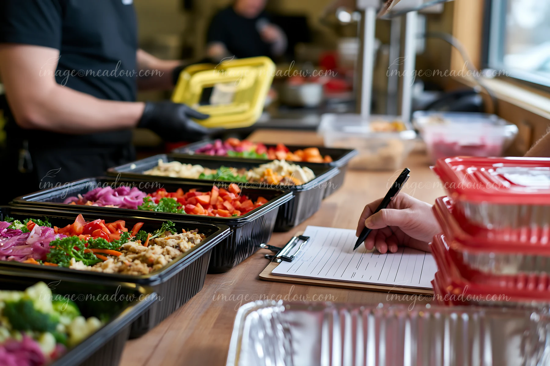 Gloved worker packs buffet food into containers beside a clipboard and prepared dishes.
