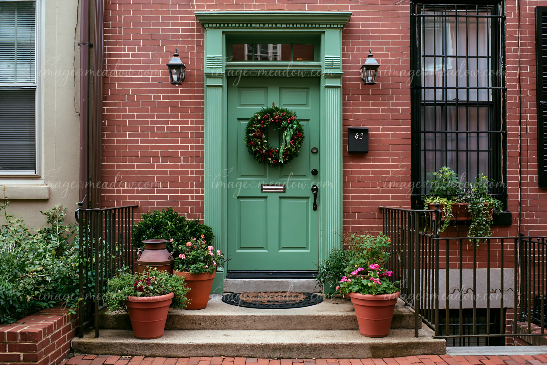 Green front door with a wreath and welcome mat at a brick townhouse entrance with potted plants.