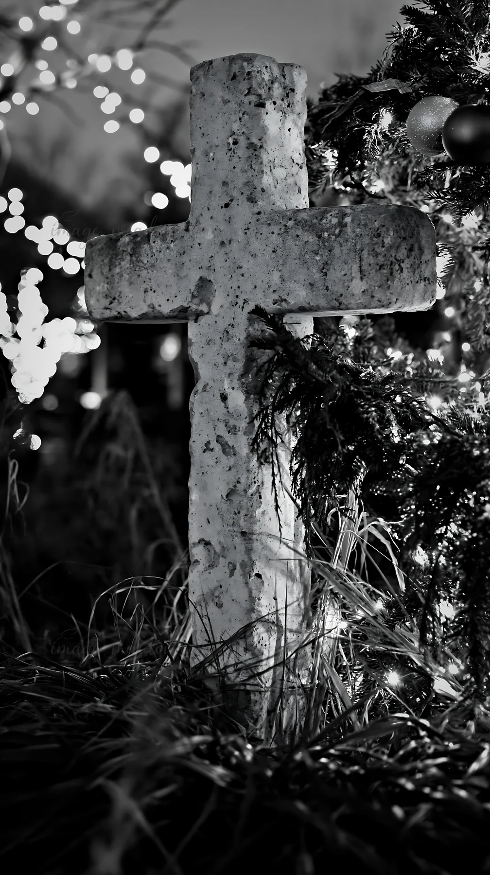 A handcrafted Christian cross stands among grass in a Christmas meadow. The scene is lit by decorative lights and surrounded by greenery, creating a festive atm