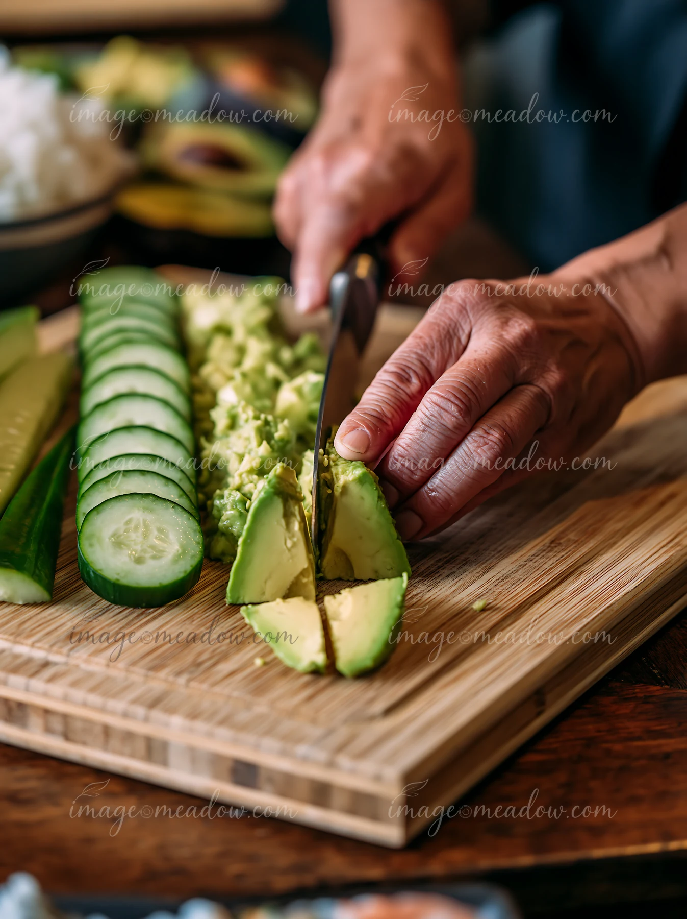 Hands are busy slicing avocado and cucumber on a wooden board.