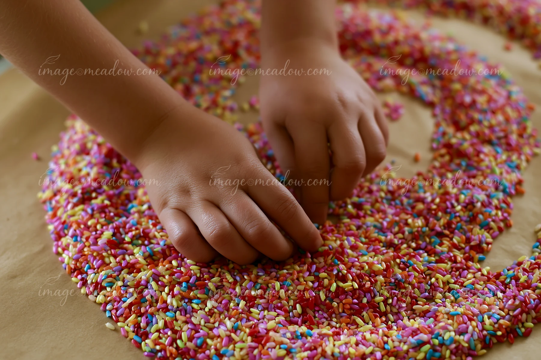 Hands arrange multicolor rice grains forming swirl on a flat surface.