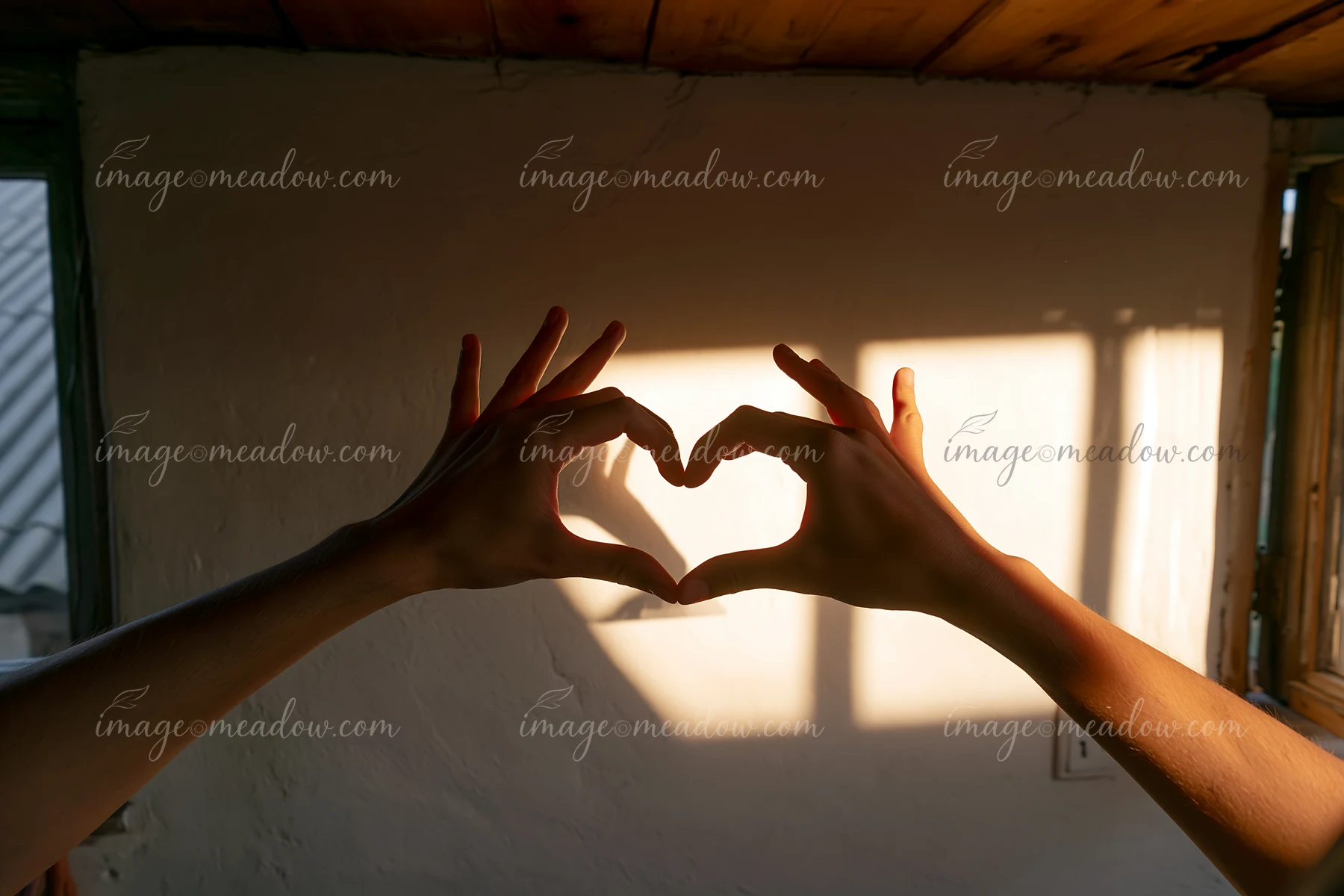 Hands create a heart shape casting a shadow on a wall in a warm room with wooden features.